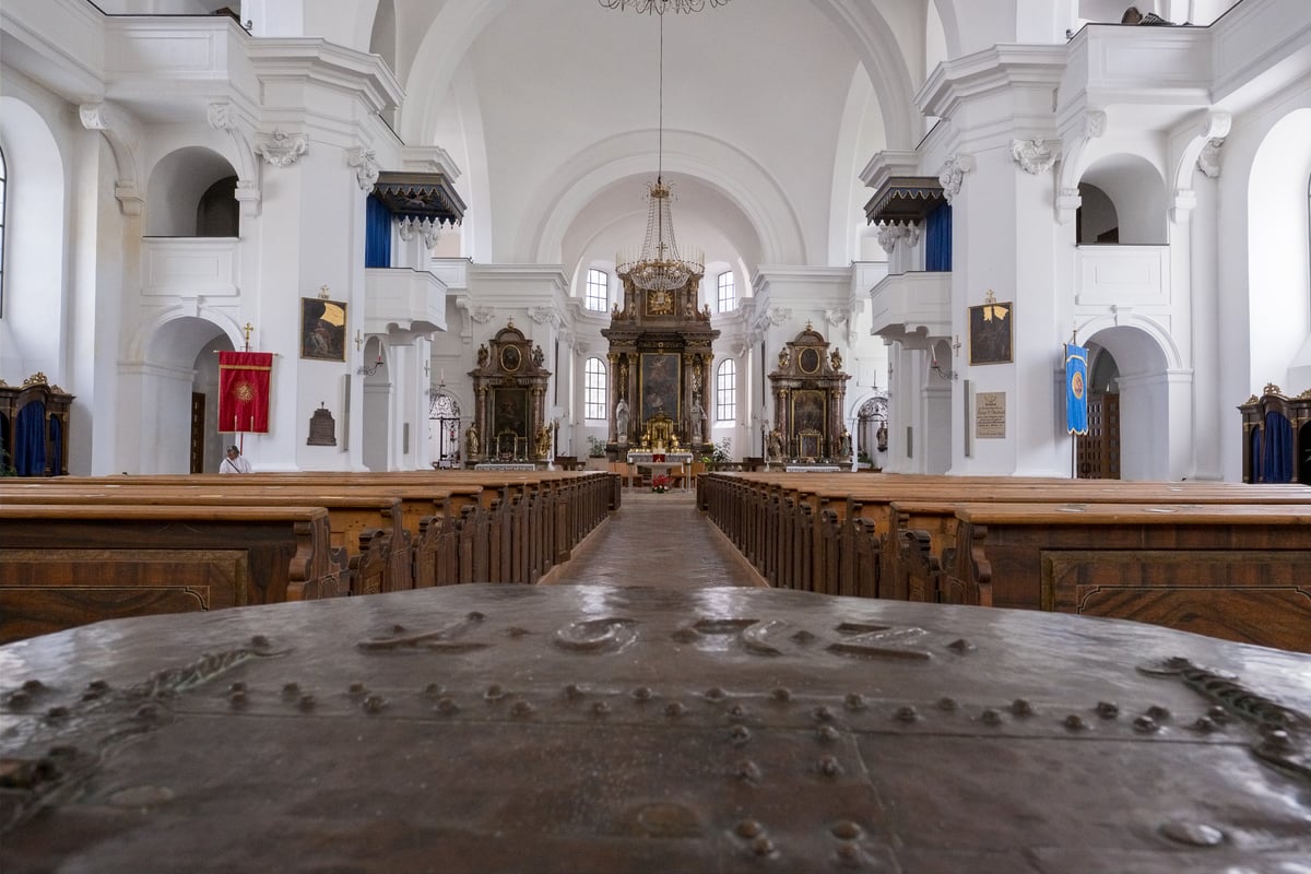 The white baroque nave of Hallein parish church seen down the central aisle from behind the studded metal lid of the baptismal font, with the gilded high altar at the far end.