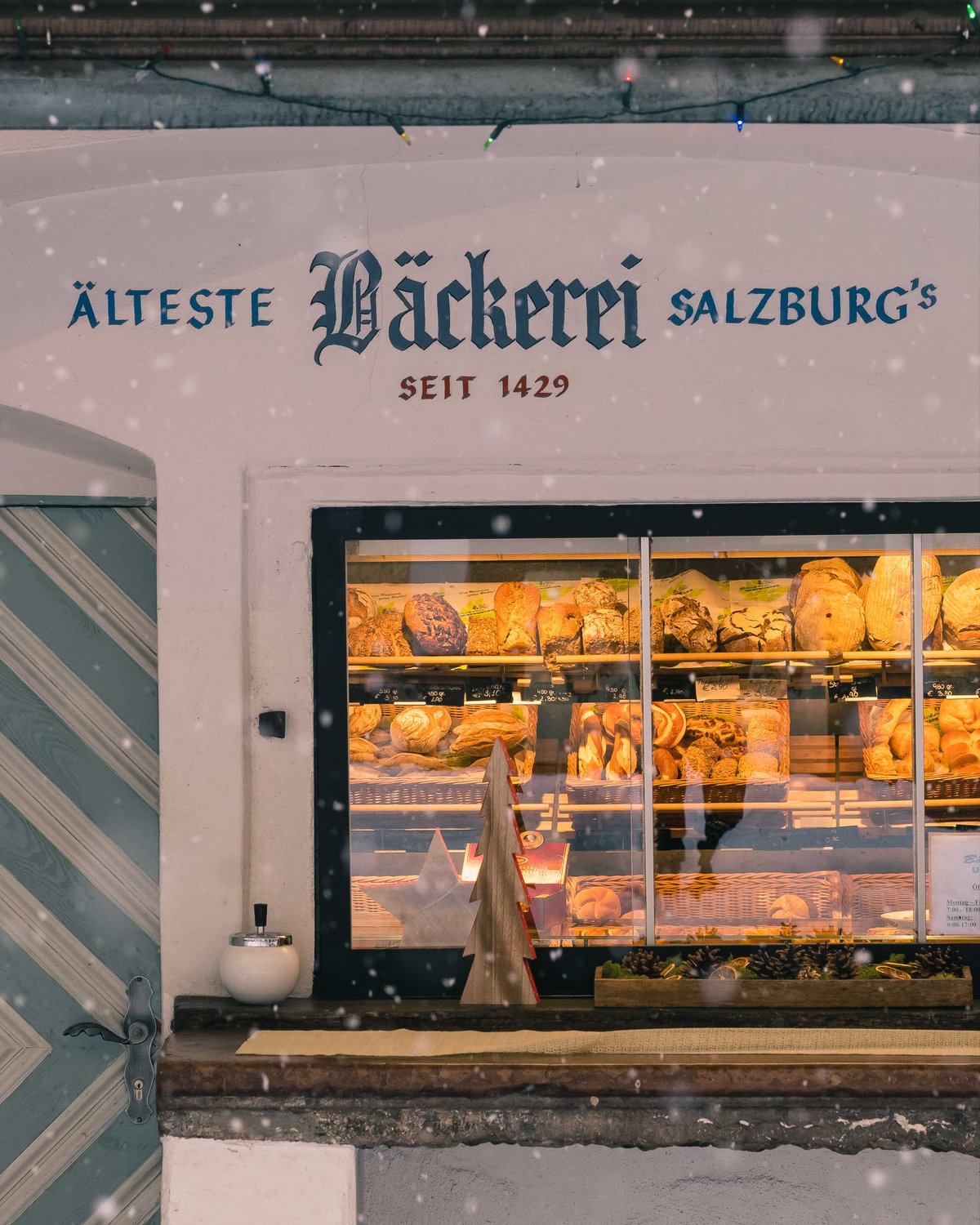 Snow falls past a Salzburg bakery storefront with Aelteste Baeckerei Salzburg's lettering above in winter