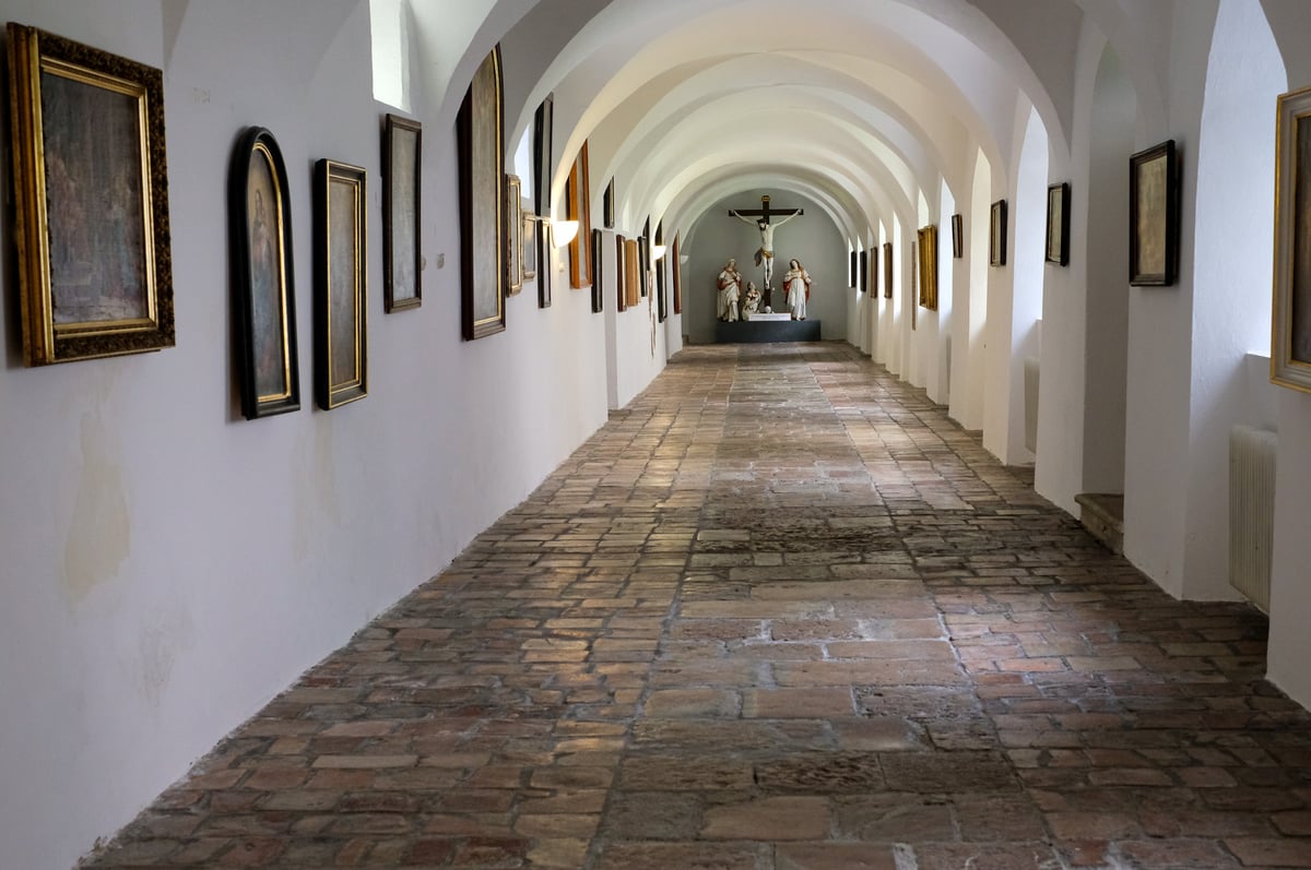 A long vaulted whitewashed corridor at the Mattsee collegiate foundation, hung with old framed paintings and closed at the far end by a carved crucifixion group in a niche.