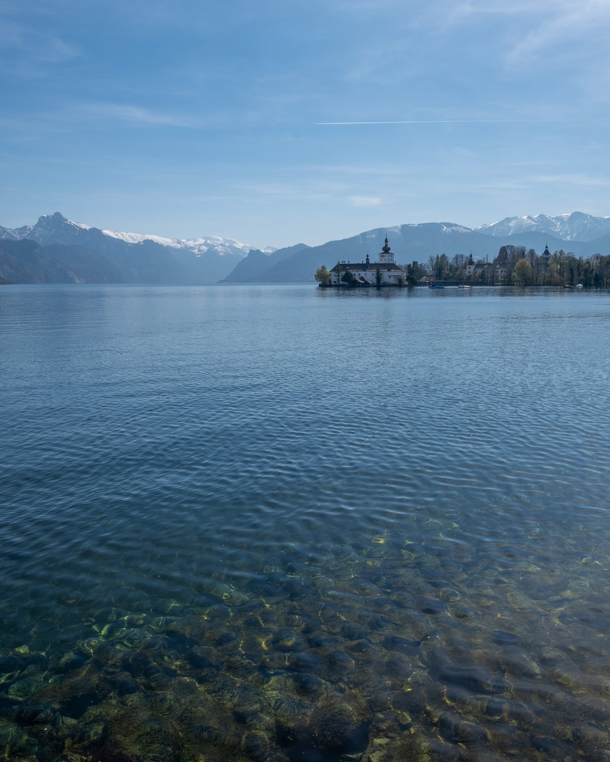 The Traunsee seen across clear stony shallows, with Seeschloss Ort on its island to the right and snow-capped Salzkammergut peaks on the far shore.