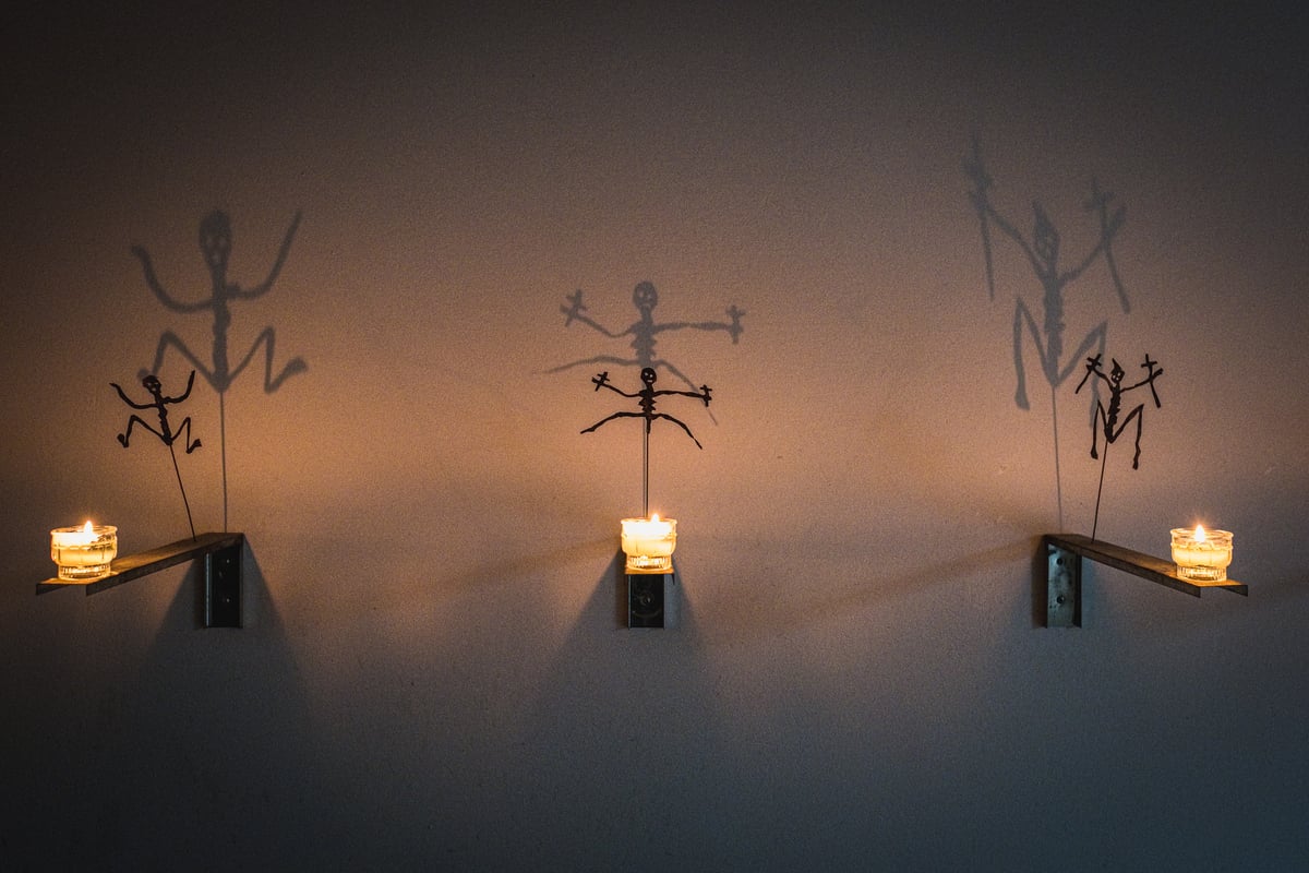 Three candles on wall brackets each lift a cut-metal skeleton figure whose shadow spreads large and dancing across the crypt wall beneath Salzburg Cathedral.