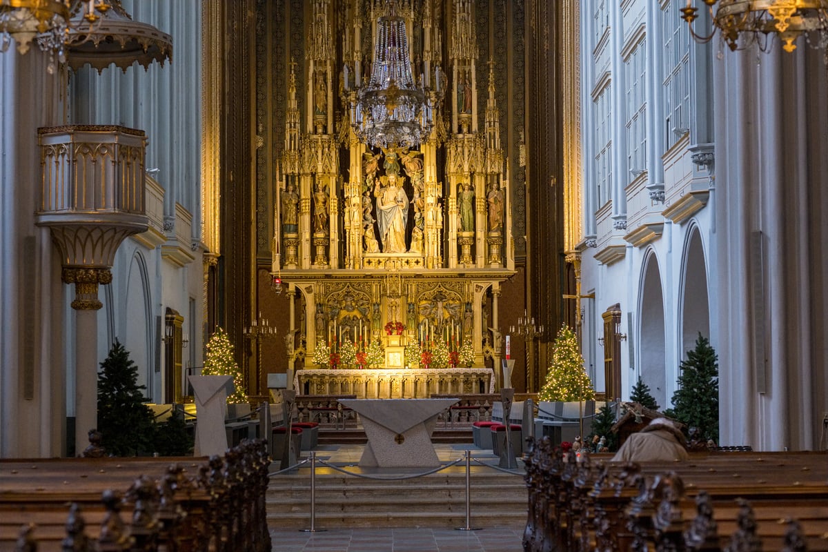 A towering gilded neo-Gothic high altar with a blessing figure of Christ holding an orb glows at the end of a white columned nave in a Vienna church, flanked by lit Christmas trees and a crib.