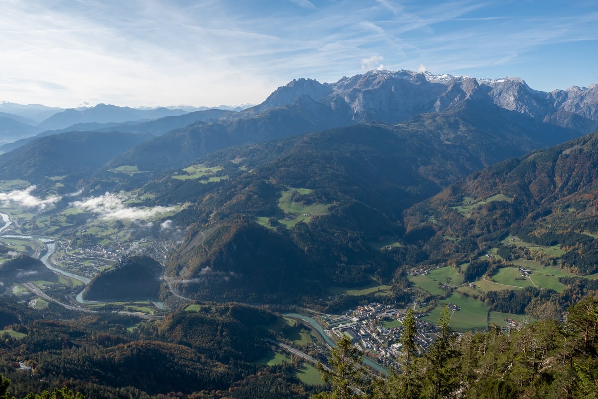 A high panorama over the Salzach valley with the town of Werfen and its fortress on a rock at the left, a village with an industrial plant along the turquoise river below, green meadows and forest on the slopes, and a snow dusted limestone massif on the horizon.