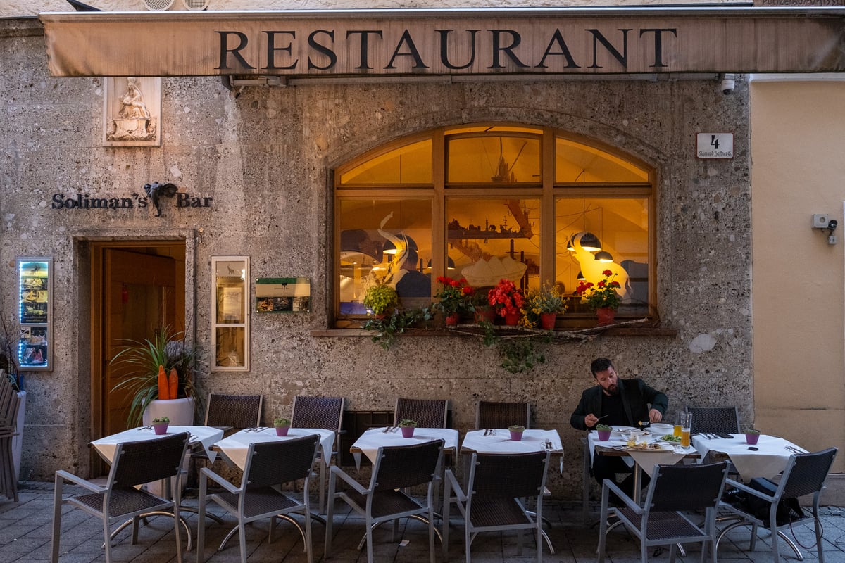 The conglomerate stone facade of the Hotel Elefant building under a Restaurant awning, with a Soliman's Bar sign, an arched window full of warm light and elephant murals, geraniums on the sill and a single diner at the row of outdoor tables.