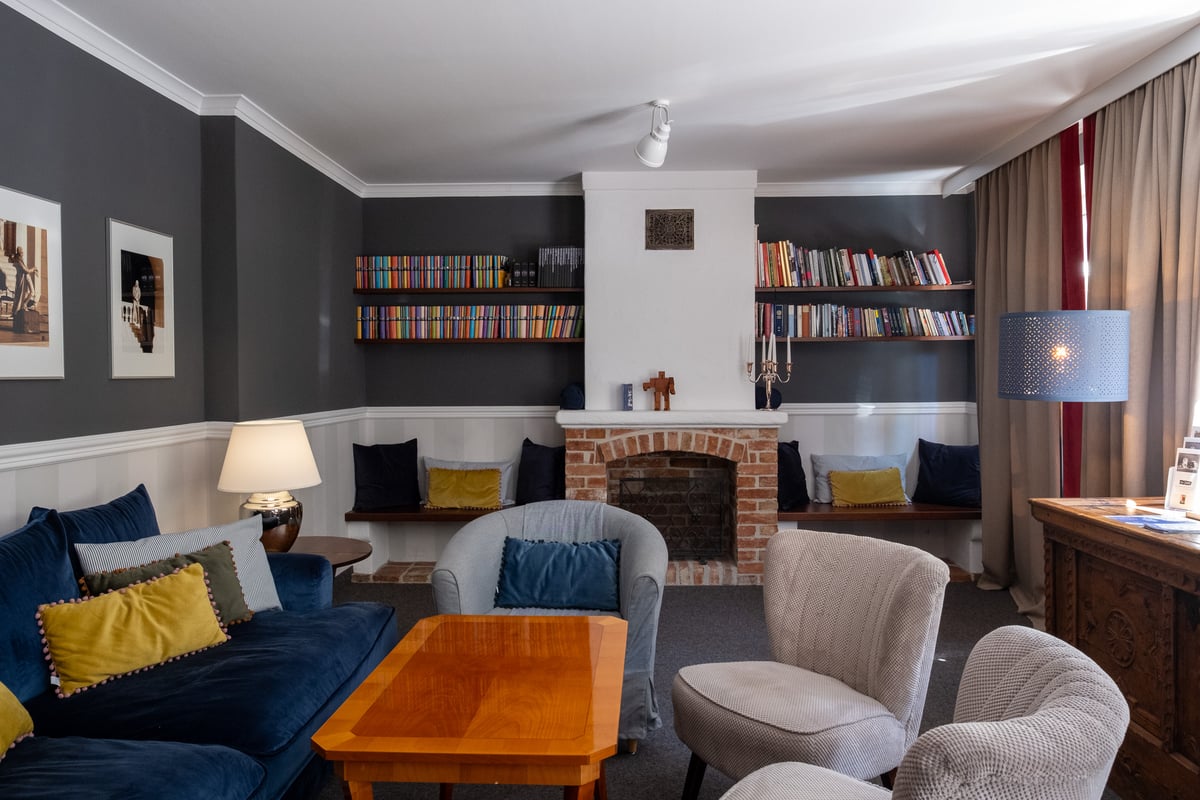 A hotel lounge in Salzburg with a whitewashed chimney breast over a brick fireplace, long shelves of colour-sorted books, a deep blue velvet sofa and cushioned window benches.