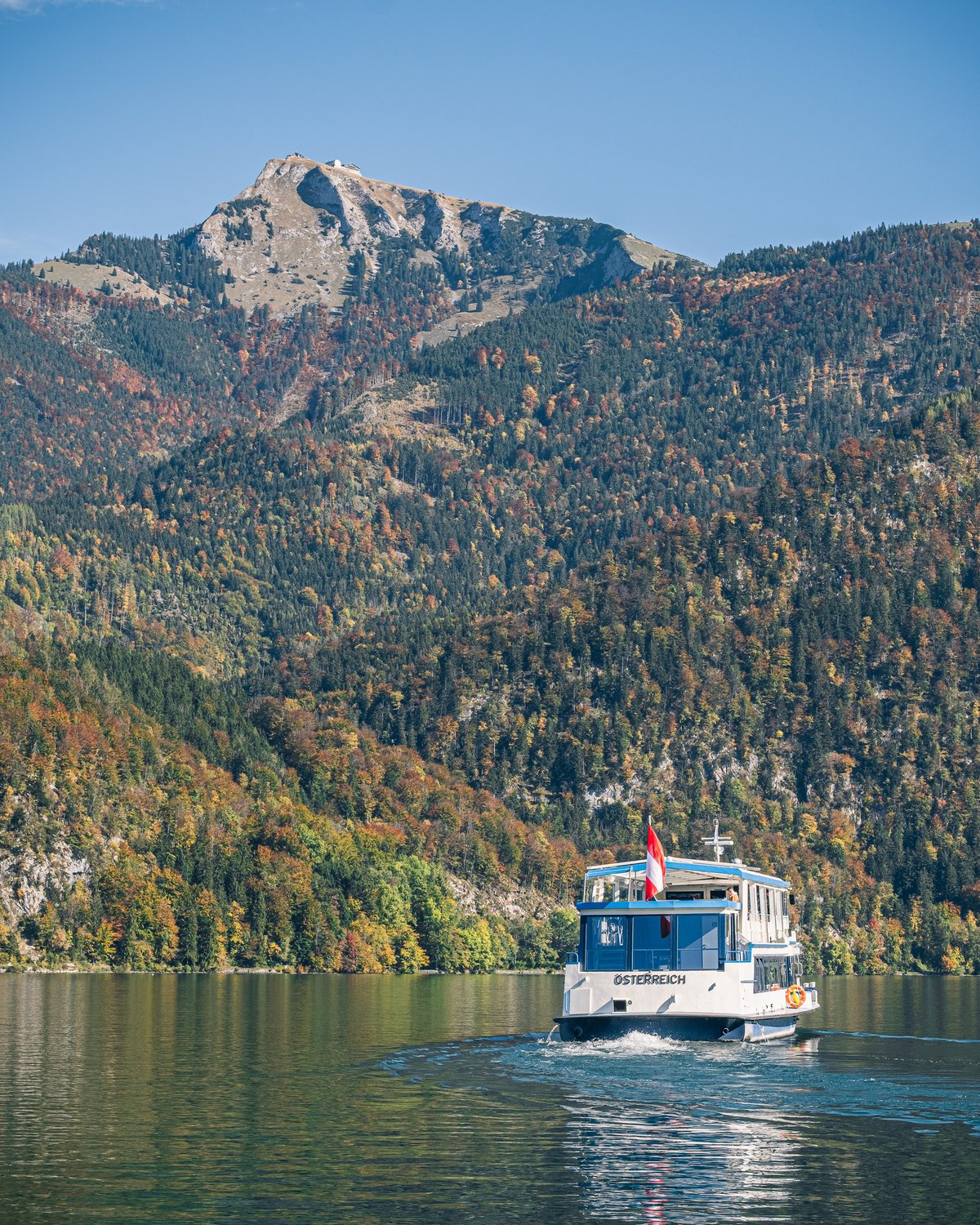 The ferry MS Österreich crosses the Wolfgangsee below the Schafberg, whose bare rock summit rises above slopes of red and gold autumn forest.