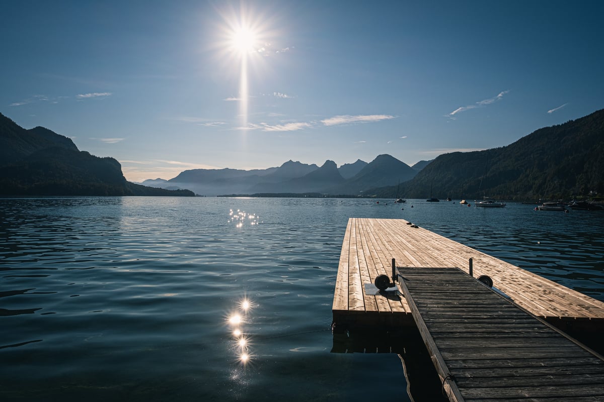 A pale wooden jetty runs out into a calm Wolfgangsee under a high autumn sun that throws a starburst on the sky and glints on the water, with moored sailing boats to the right and layered peaks closing the far end of the lake.