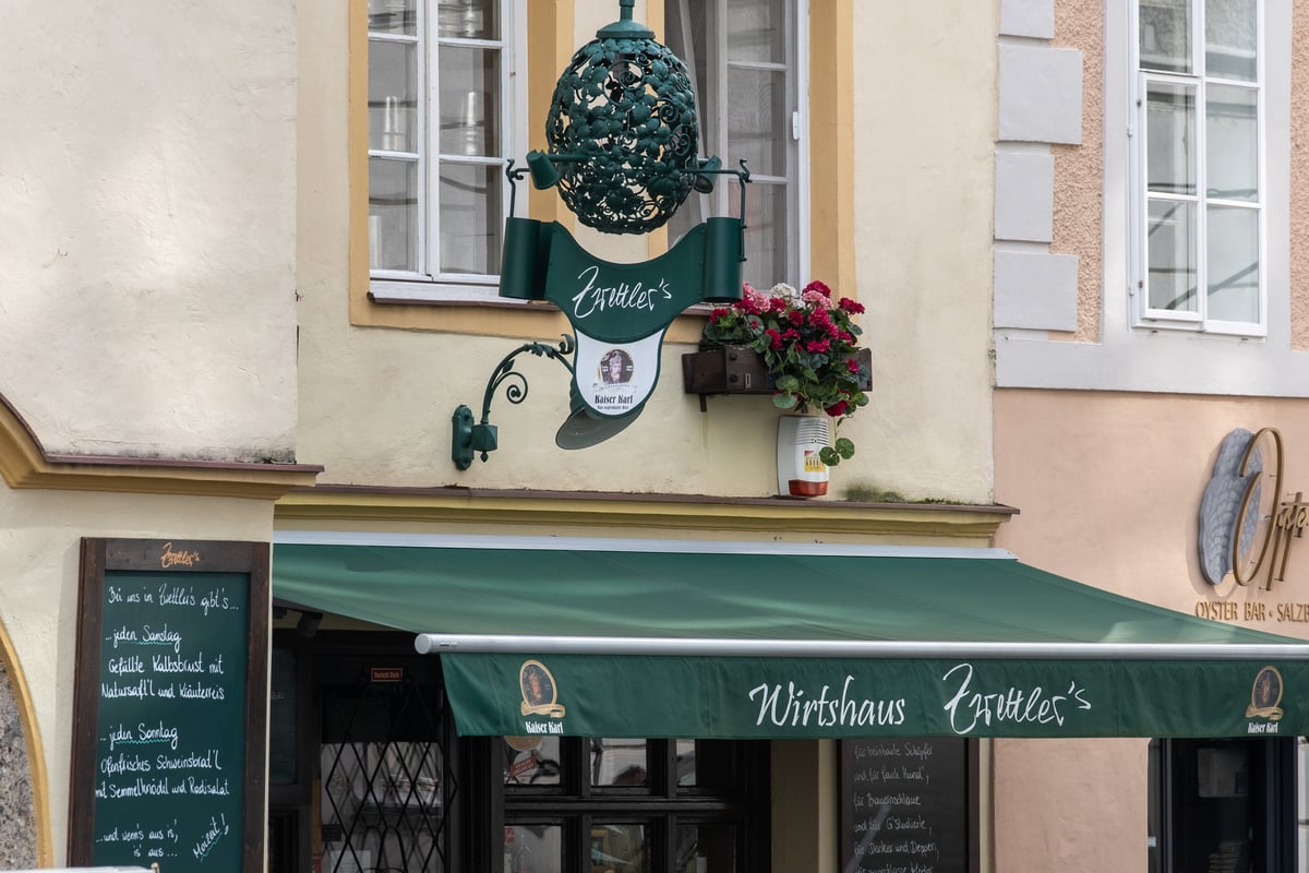 A green wrought-iron guild sign lettered Zwettler's hangs beside a geranium box above the green Wirtshaus Zwettler's awning in Salzburg, with a handwritten menu board at the left.