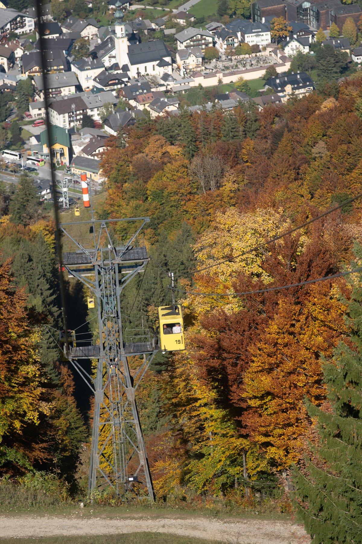 A yellow single cabin of the old Zwölferhorn cable car passes a lattice pylon above autumn beech forest, with the village of St. Gilgen and its church spread out in the valley below.