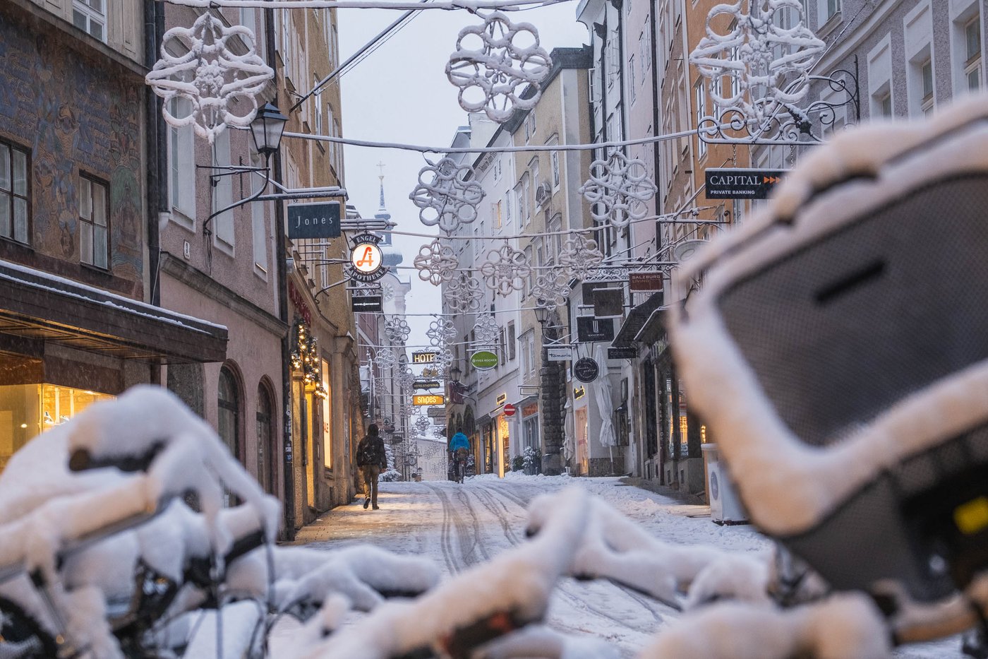 Christmas decorations hang above Getreidegasse with snow-covered bikes and Rathausturm in the distance in Salzburg old town.