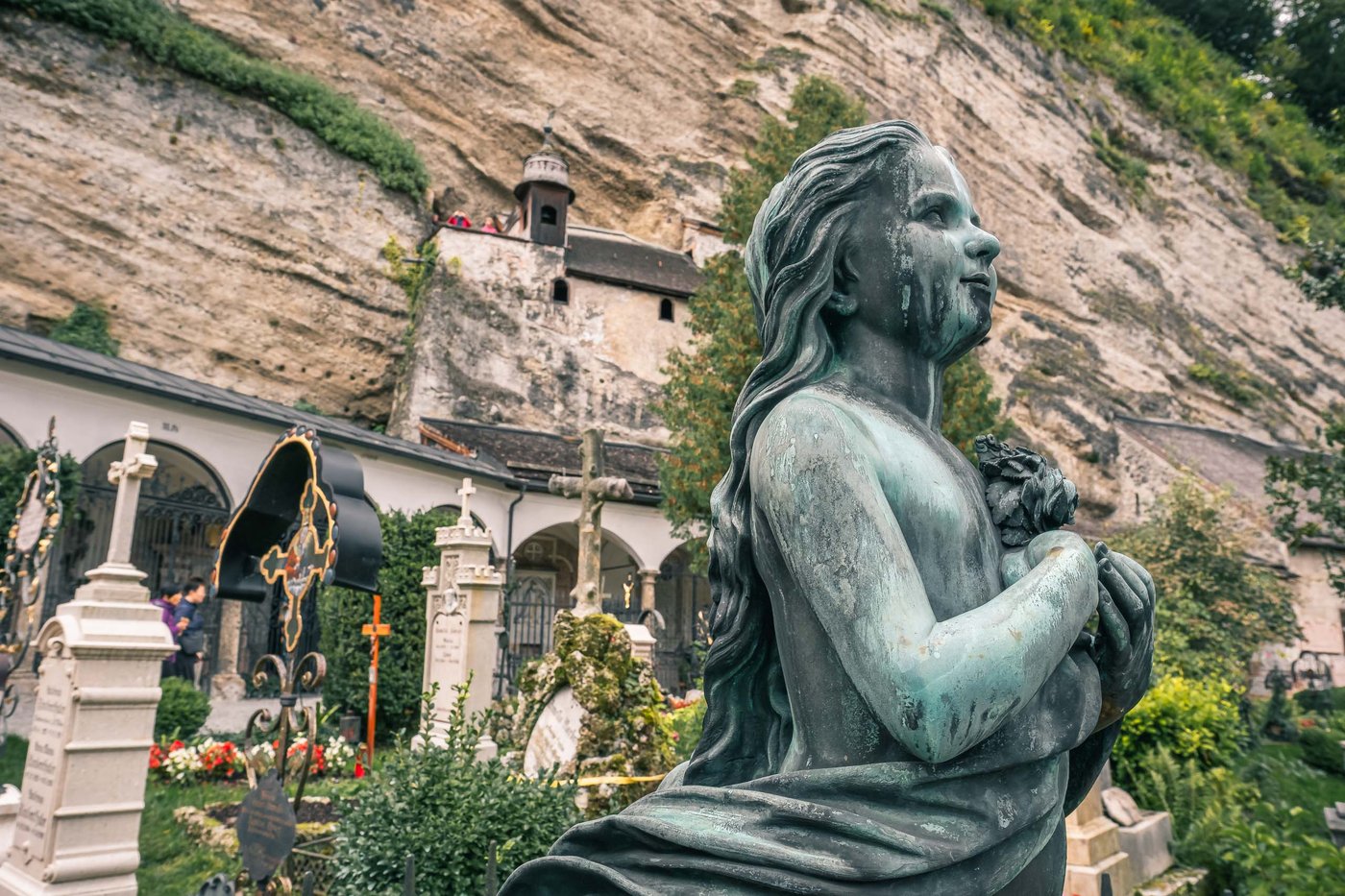 Bronze angel grave statue with catacomb chapels in the cliff behind at St Peter's Cemetery in Salzburg.