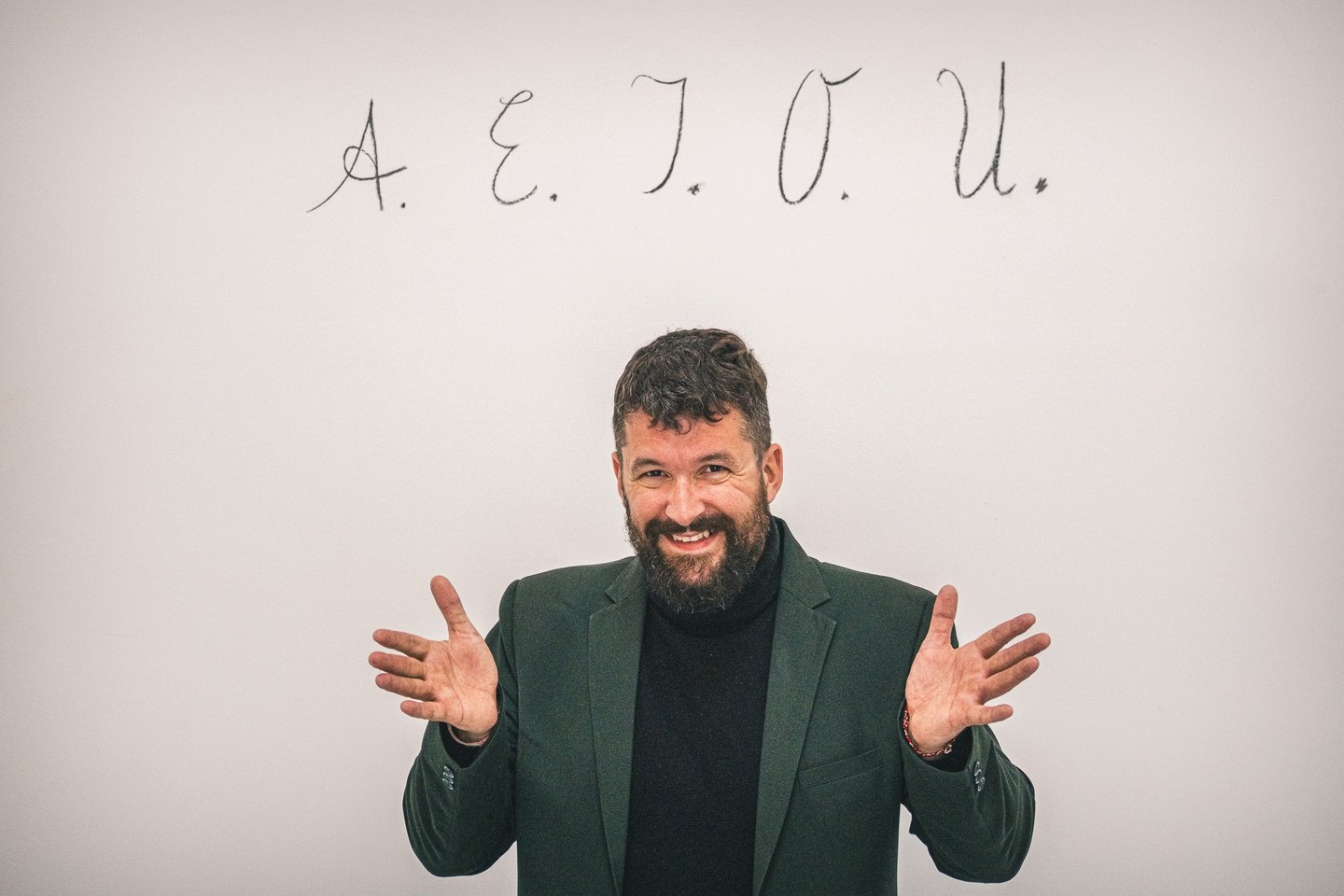A smiling bearded man in a dark green blazer and black turtleneck spreads both hands wide, standing beneath the letters A. E. I. O. U. written in cursive on a pale wall.