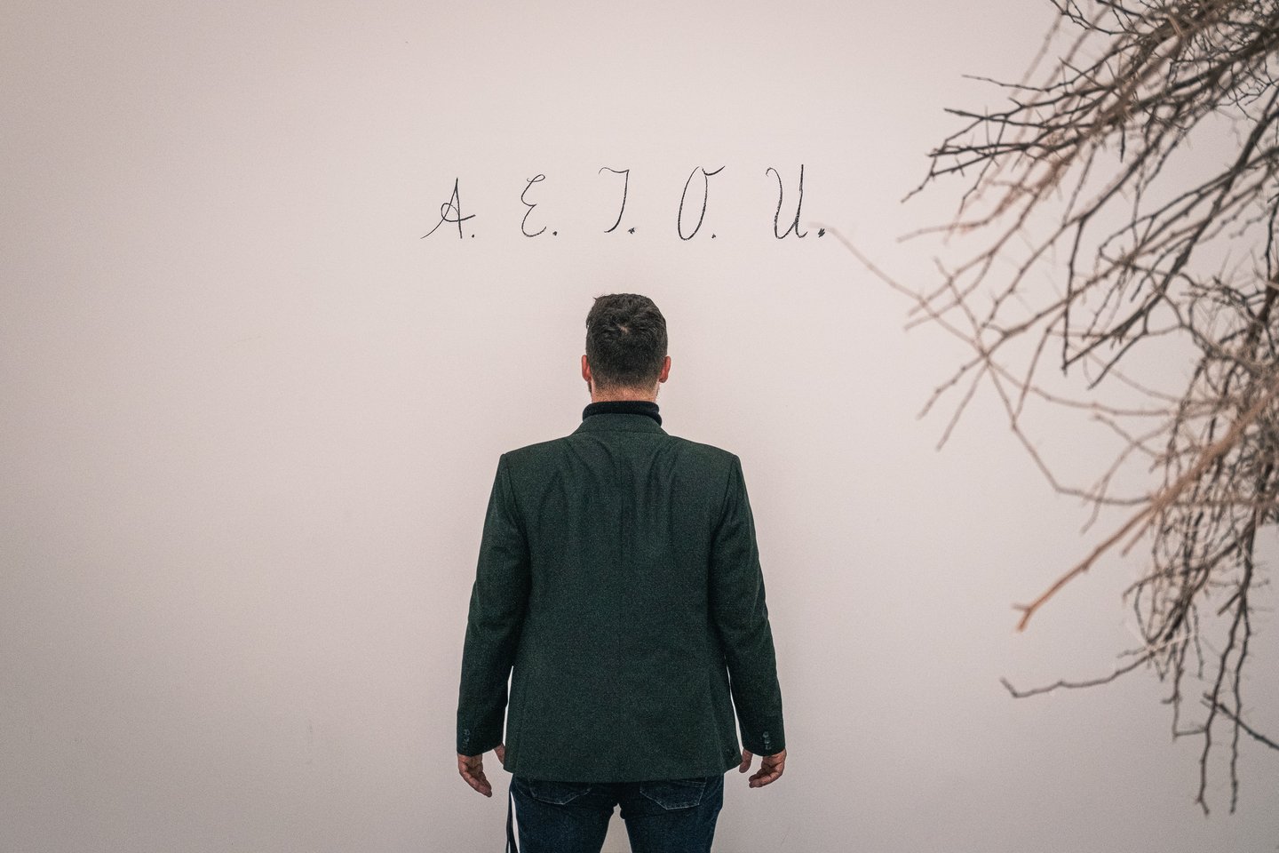 A man in a dark green jacket stands with his back to the camera facing the letters A. E. I. O. U. written in thin cursive across a pale wall, with dry bare branches hanging into the frame on the right.
