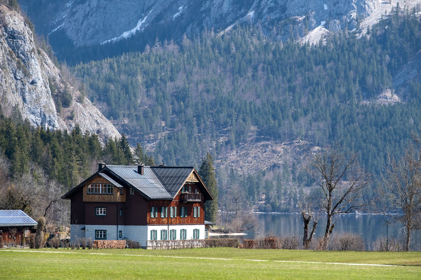 A dark-timbered Ausseerland house with green shutters stands on a green meadow by the shore of the Altausseer See, under a forested slope and bare limestone cliffs in early spring.