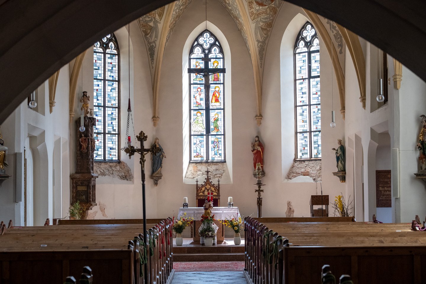 The white vaulted interior of the parish church in Altaussee, looking down the wooden pews to the altar beneath three tall stained glass windows.