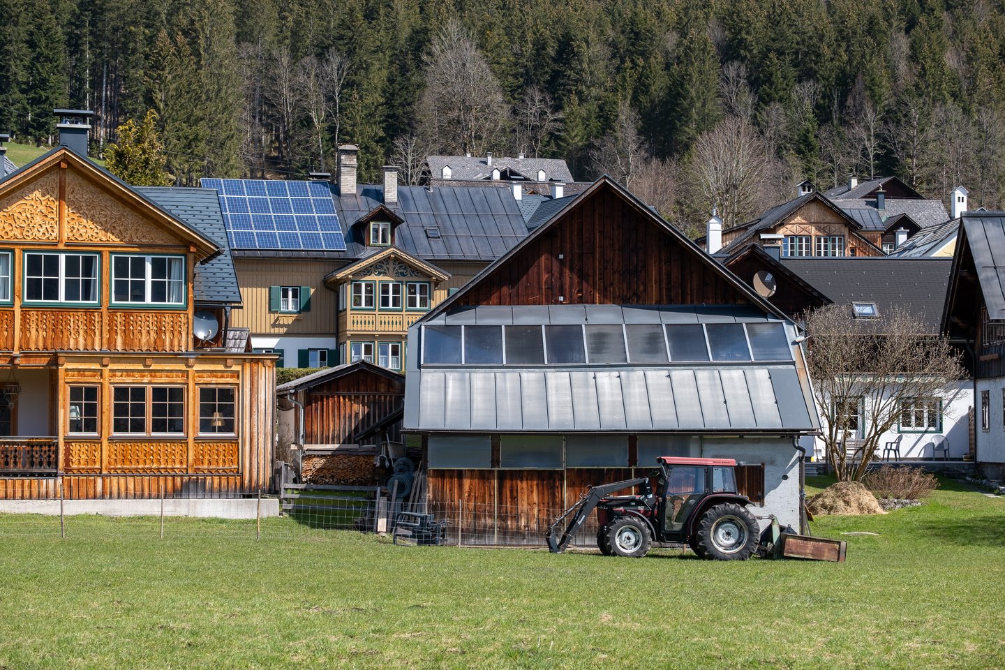 Carved wooden Ausseerland houses and a hay barn with a red tractor parked beside it, on a green spring meadow below a forested slope in Altaussee.