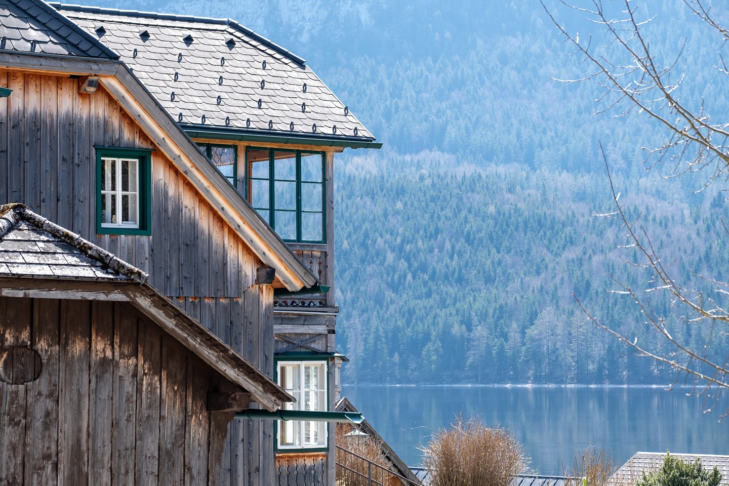 A weathered wooden Ausseerland house with a shingled roof, green window frames and a glazed corner gallery stands above the Altausseer See, with the forested far shore behind it.