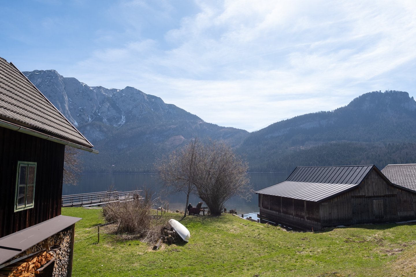 A dark wooden house with a stacked woodpile and a boathouse stand on a grass slope at the edge of the Altausseer See, with rock faces and forested ridges rising beyond the far shore.