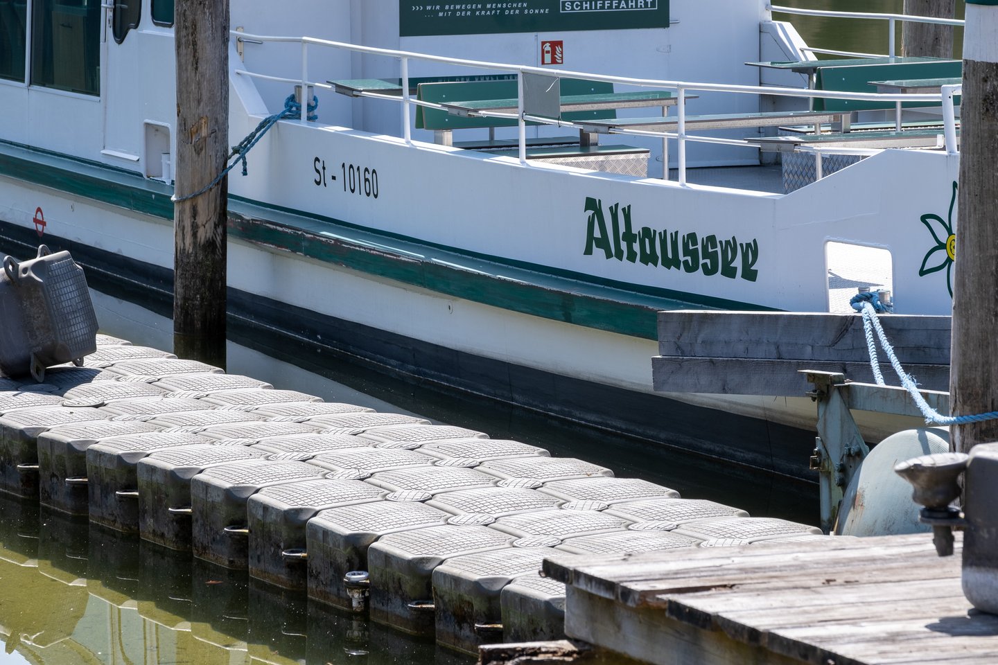 The excursion boat Altaussee, registered St-10160, lies moored against a floating pontoon dock on the Altausseer See, its green benches empty.