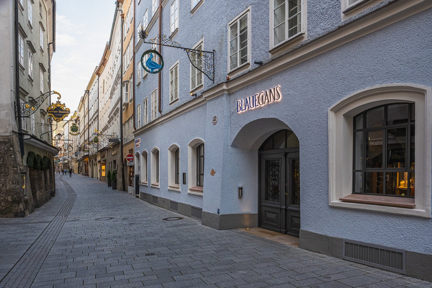 The pale blue facade of the Arthotel Blaue Gans on Getreidegasse at dusk, with its illuminated BLAUEGANS lettering above the arched entrance and the blue goose guild sign on a wrought-iron bracket.