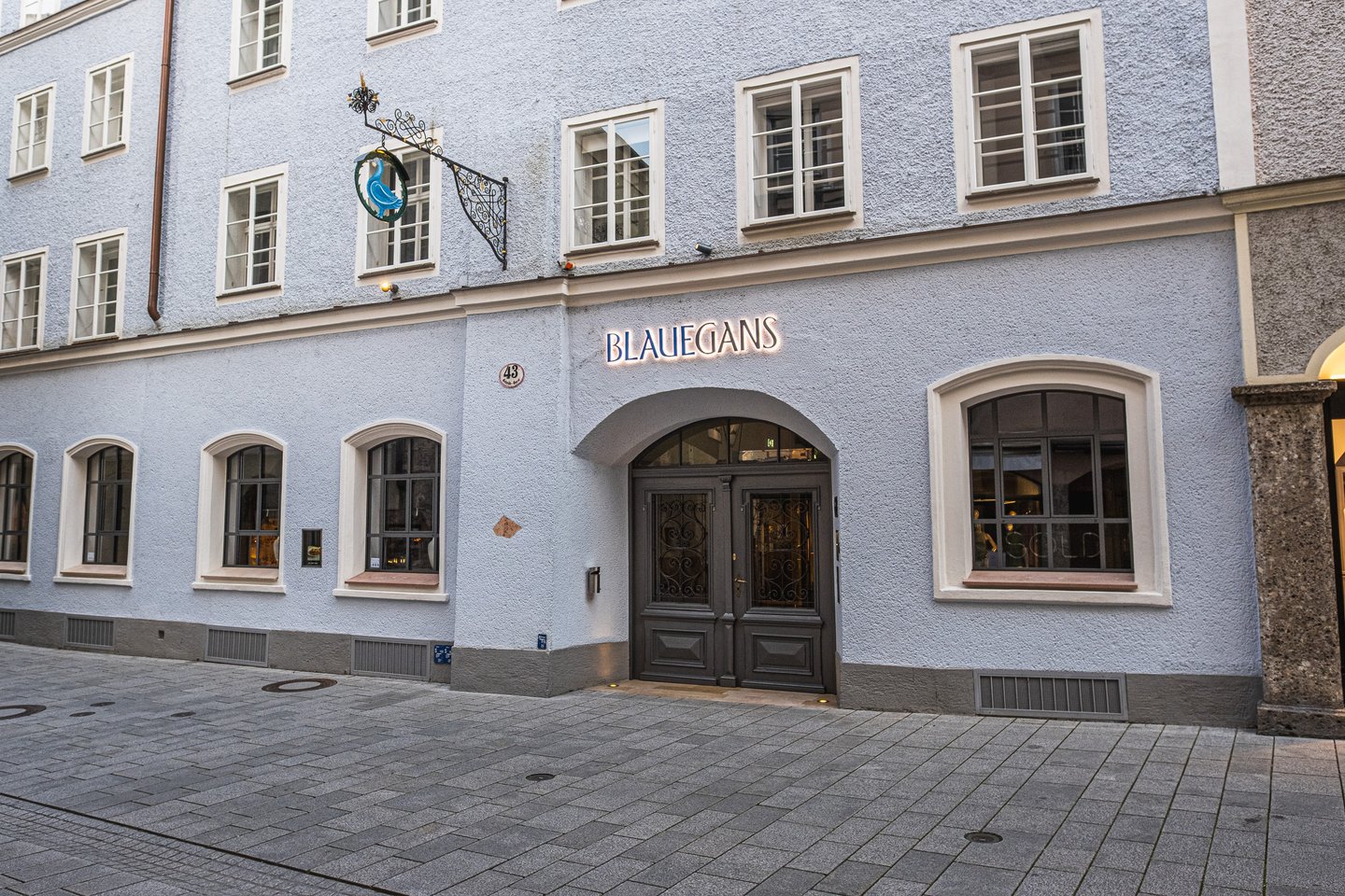 The Getreidegasse entrance of the Arthotel Blaue Gans in Salzburg, a pale blue rendered facade at number 43 with a wrought-iron guild sign showing a blue goose, illuminated BLAUEGANS lettering above the arched double doors and lit restaurant windows either side.
