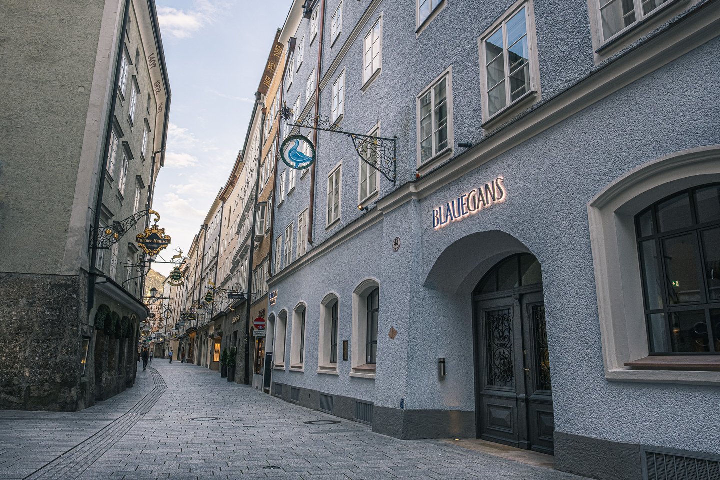 Looking steeply up the blue-grey facade of the Arthotel Blaue Gans on an almost empty Getreidegasse, with the goose guild sign and the lit BLAUEGANS lettering above the dark arched door.