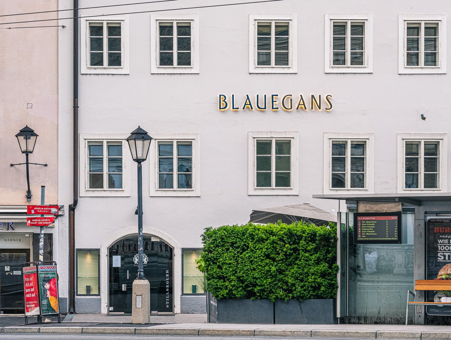 The pale facade of the Arthotel Blaue Gans on Herbert-von-Karajan-Platz in Salzburg, with gold BLAUEGANS lettering between rows of white-framed windows, a Stillsegler shopfront below, a clipped hedge and a bus stop display in the evening light.