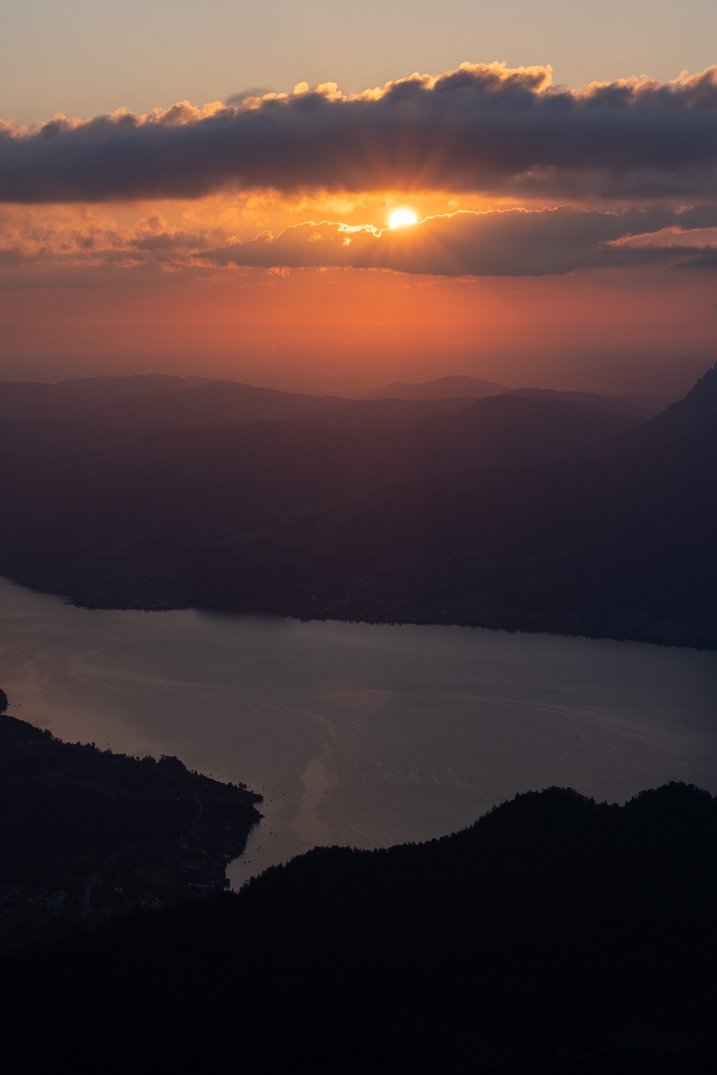 The rising sun breaks through a dark band of cloud and throws an orange path across the Attersee, seen in telephoto from the Schafberg over silhouetted ridges.
