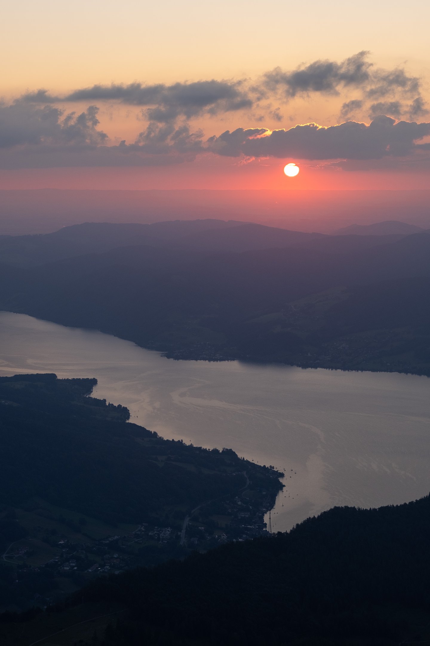 The sun clears a band of cloud above the Attersee at sunrise, seen in telephoto from the Schafberg with the long lake and its shoreline villages far below.