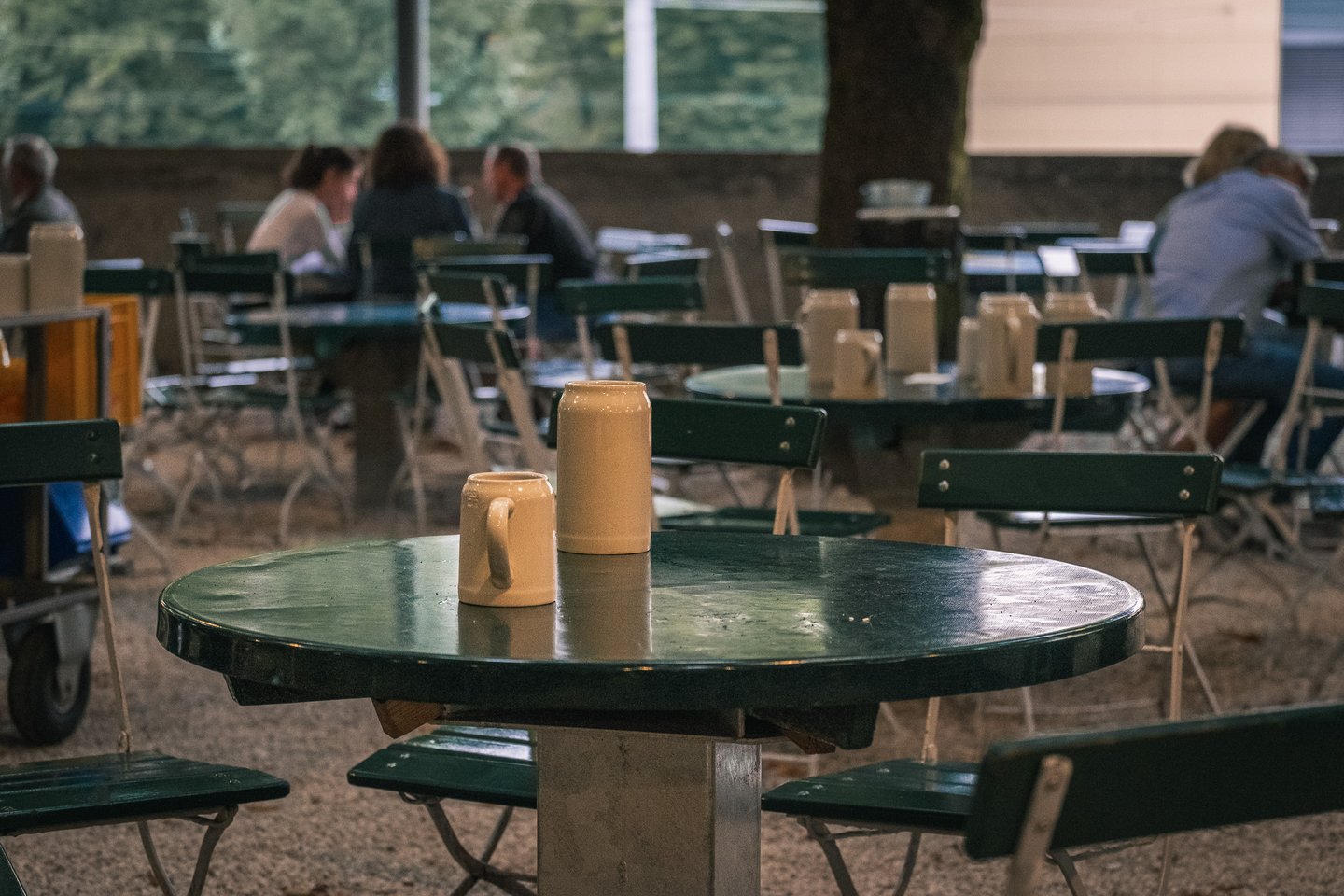Two empty stoneware mugs stand on a round green table in the gravelled chestnut beer garden of the Augustiner Braustubl, with guests talking at tables behind in the dusk.