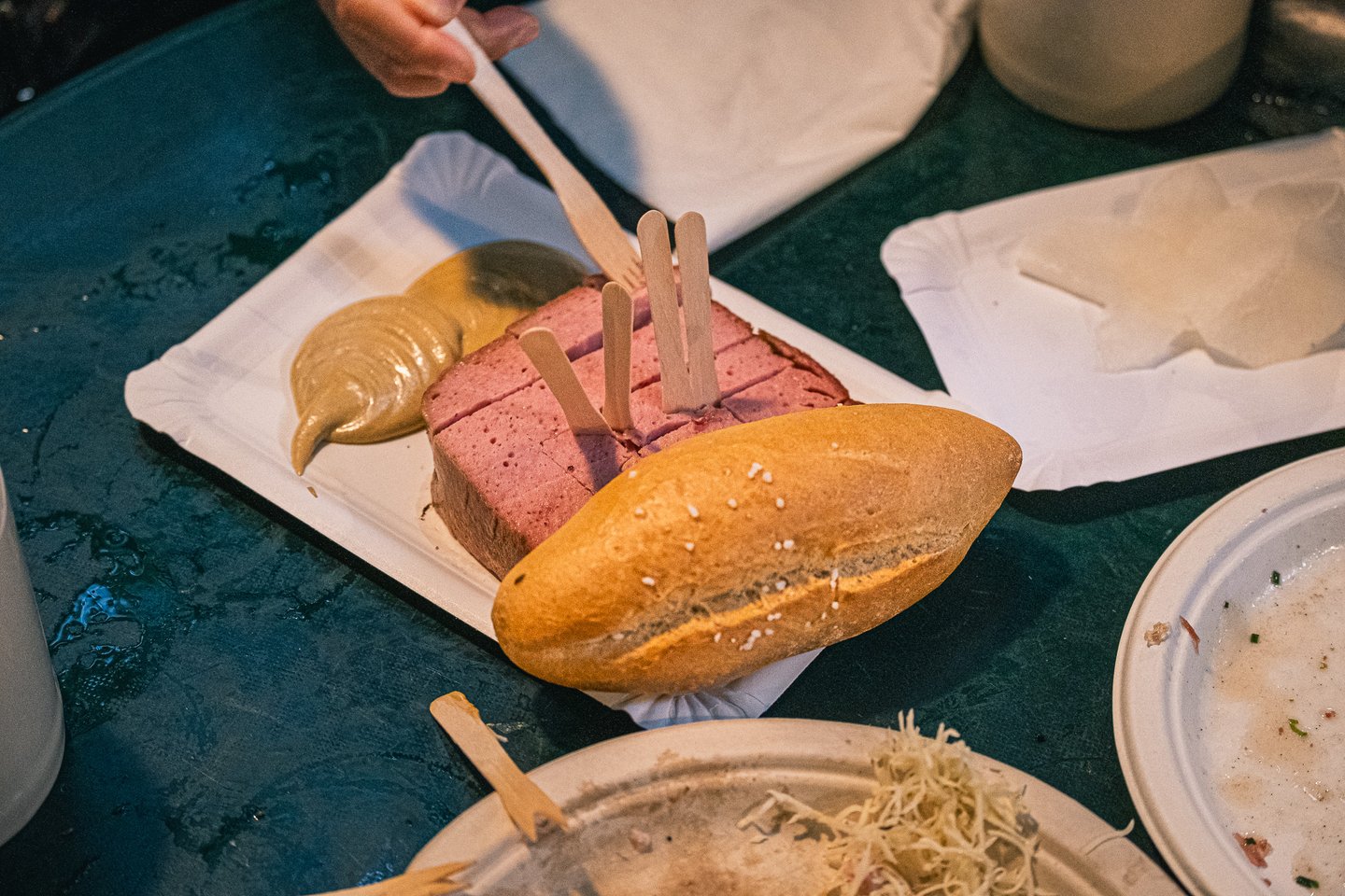 A thick slab of Leberkase, cut into squares and studded with wooden forks, lies beside two scoops of mustard and a salted white roll on a green table at the Augustiner Braustubl.