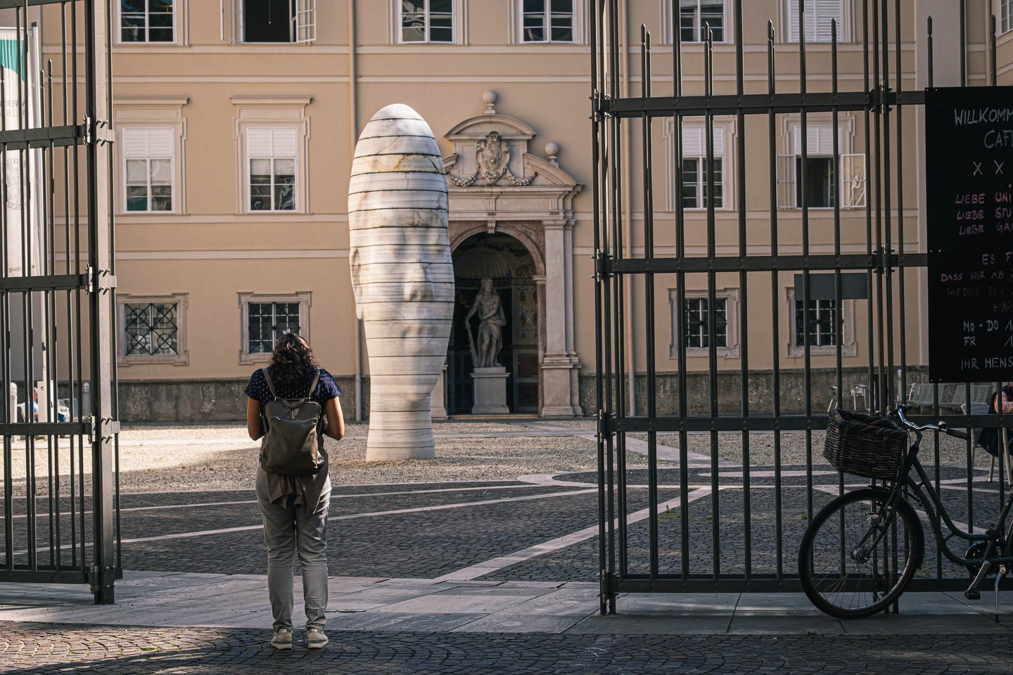 Tall white Awilda sculpture stands in a stone courtyard beyond an iron gate in Salzburg.