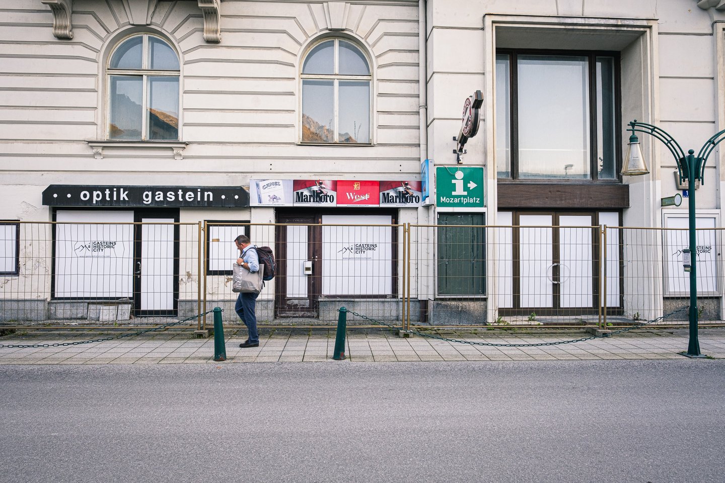 A man with a backpack walks past a row of boarded-up shopfronts in Bad Gastein, an old optik gastein sign and faded cigarette advertising still above the hoardings.