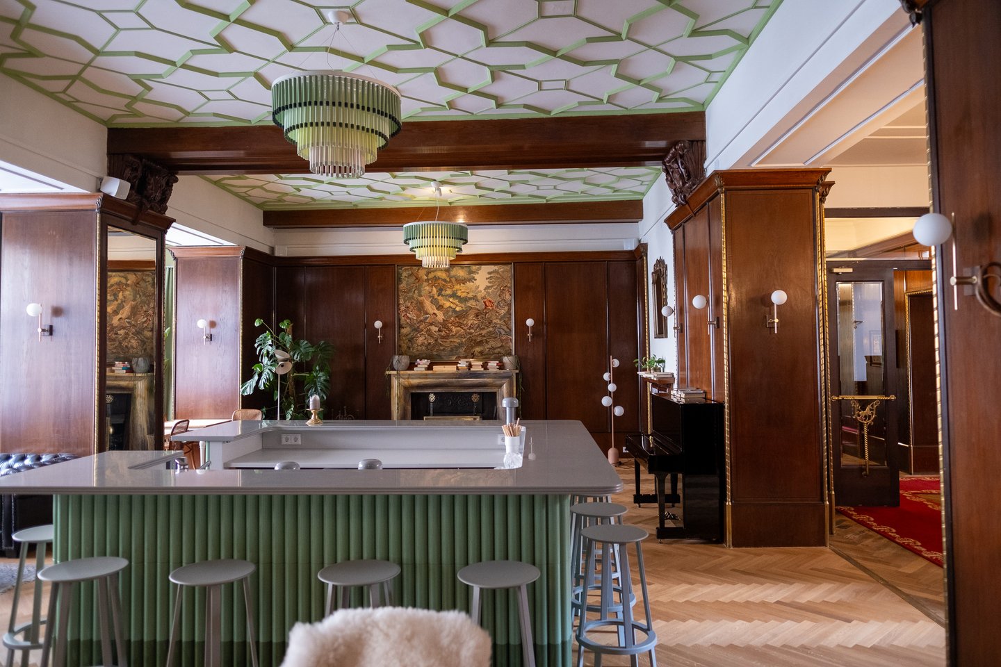 A green fluted bar counter under mint green ceiling panels in a dark wood panelled hotel lounge in Bad Gastein, with a grand piano and a tapestry above the marble fireplace.