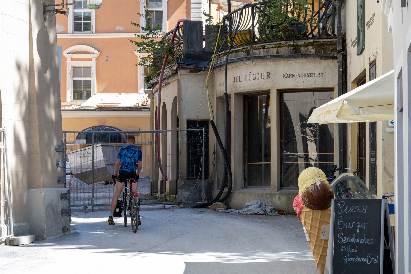 A cyclist pushes through a narrow Bad Gastein lane past construction fencing, beside a shuttered curved shopfront still lettered with an old Vienna optician's name.