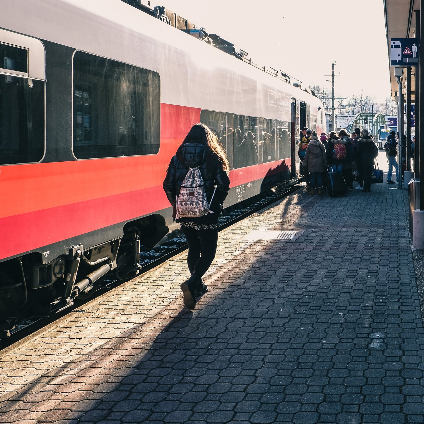 Passengers queue to board a red and white Austrian regional train on a paved platform in low winter sun, a green steel truss bridge visible beyond the station.