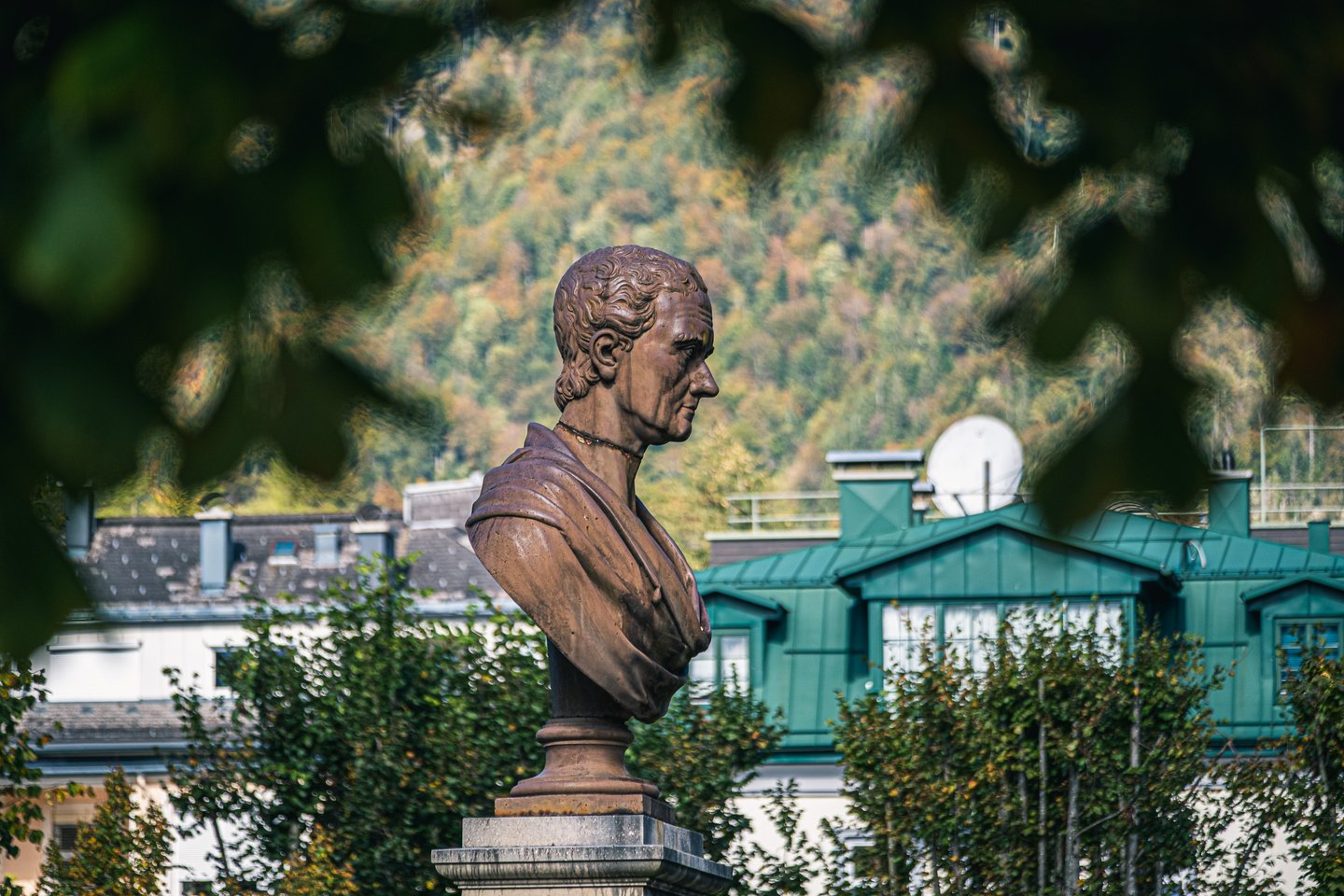 A weathered bronze portrait bust in classical drapery on a stone pedestal in a Bad Ischl park, with town roofs and a wooded hillside behind.