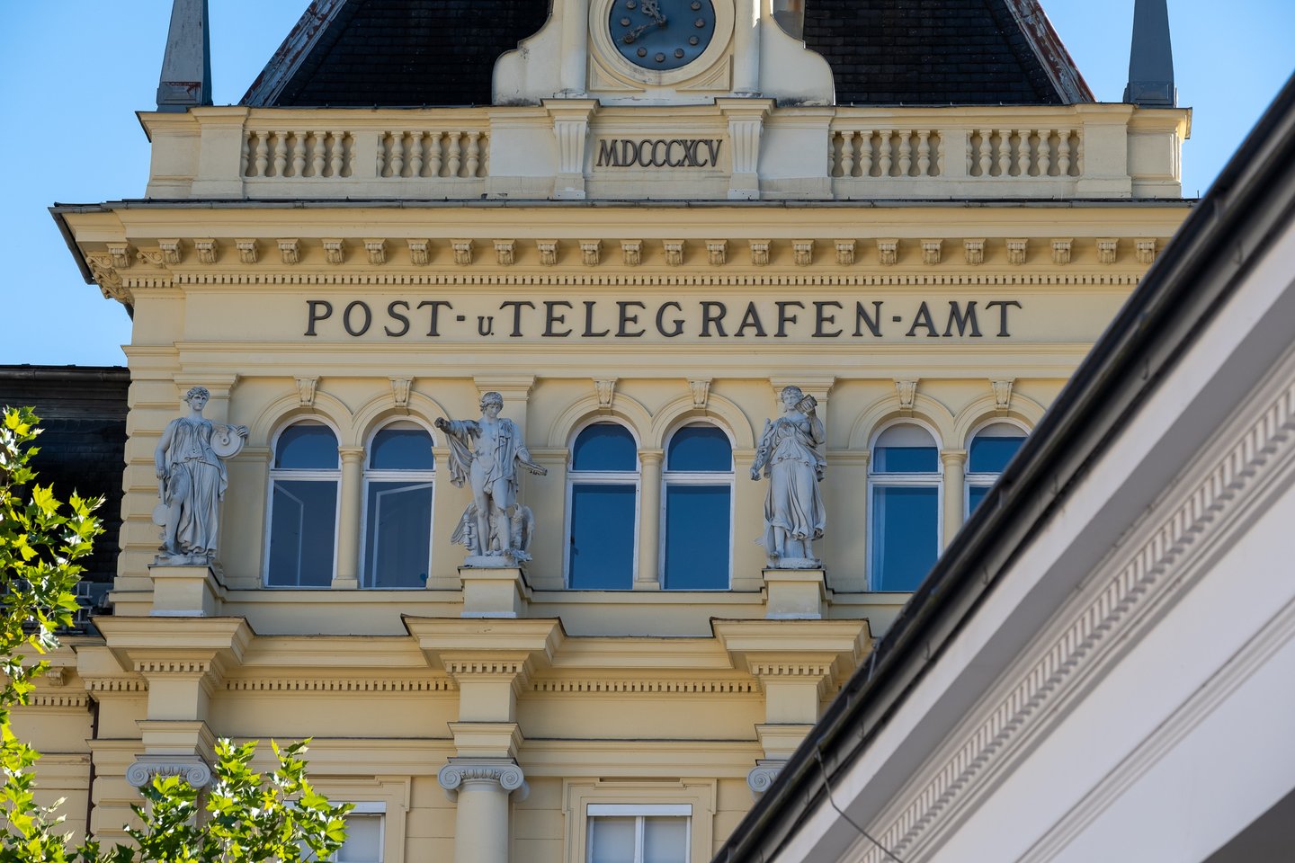 The upper facade of the old Post und Telegrafenamt in Bad Ischl, with three allegorical statues, arched windows, a clock gable and the Roman numeral date MDCCCXCV.