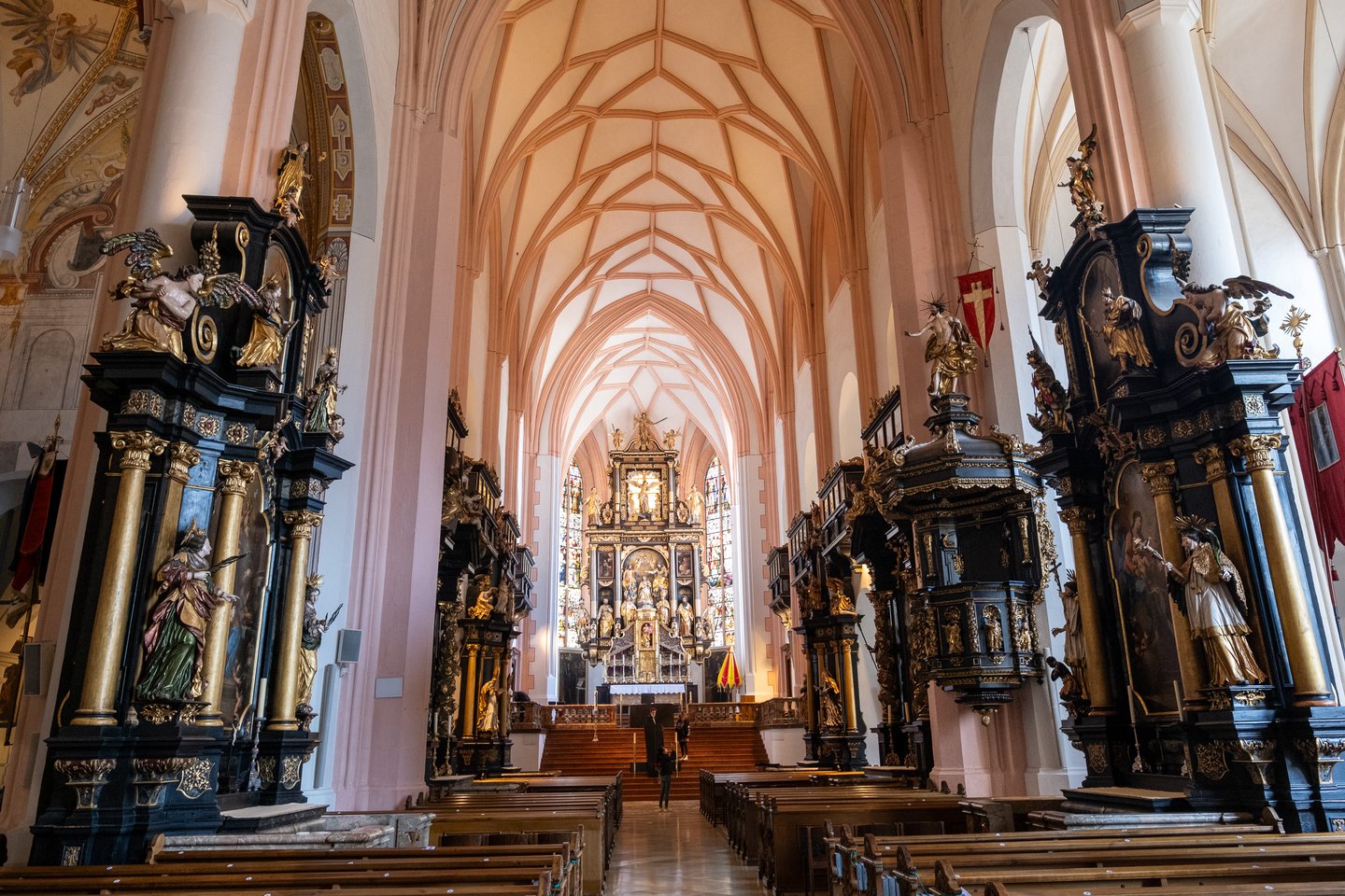The nave of the Mondsee basilica looking east toward the high altar, with the pink ribbed net vault overhead and black and gold side altars along both walls.