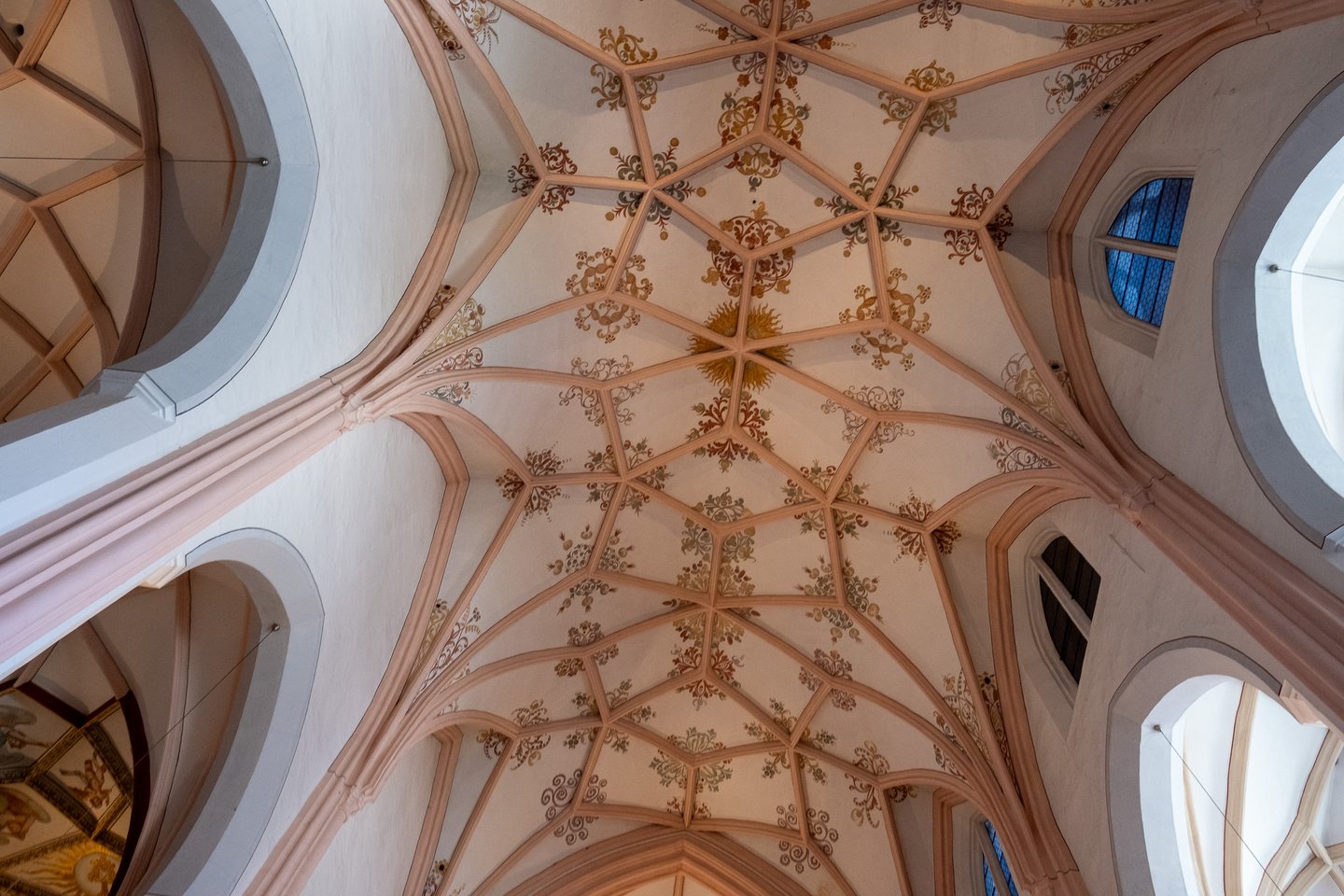 Looking straight up at the pink ribbed net vault of the Mondsee basilica, painted with gold and red tendril ornament and a sun motif at one keystone.