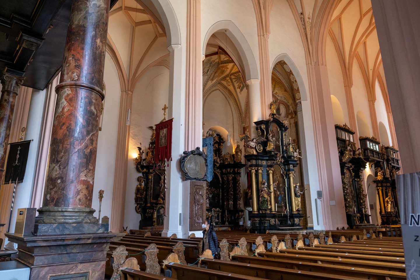 A row of black and gold side altars along the aisle of the Mondsee basilica, with a marbled column in the foreground, wooden pews and a single visitor walking past.