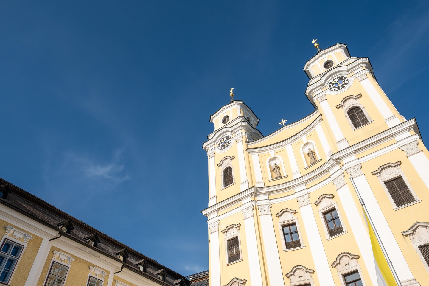 The twin towers and curved yellow gable of the Mondsee basilica seen at an angle in low evening light, with the roof of the neighbouring abbey building at the left.