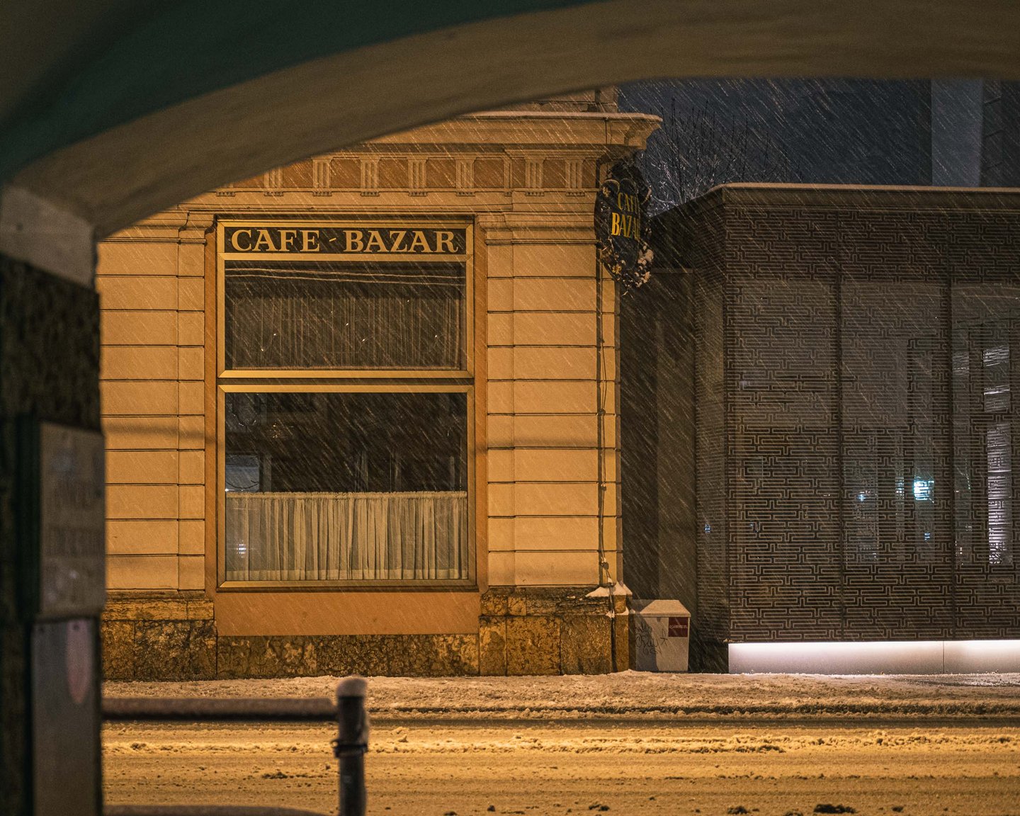 Café Bazar corner seen through an archway during heavy snow at night in Salzburg.