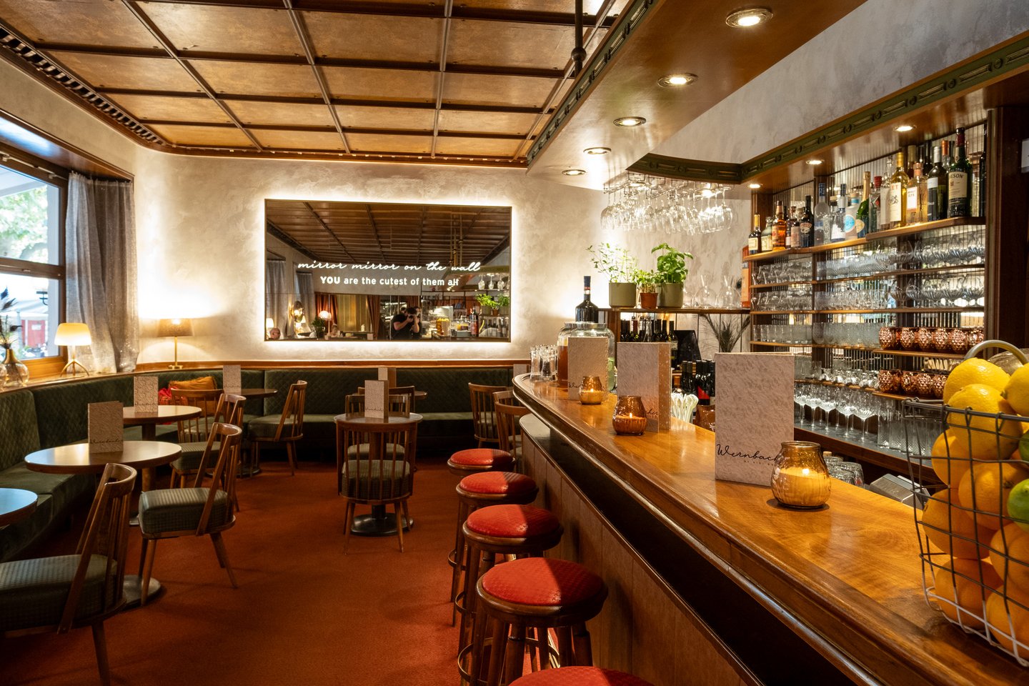 The polished wooden bar of Cafe Wernbacher runs to the right with red-topped stools, backlit shelves of glassware and spirits, herb pots and a wire basket of lemons, the green banquettes and lettered mirror to the left.