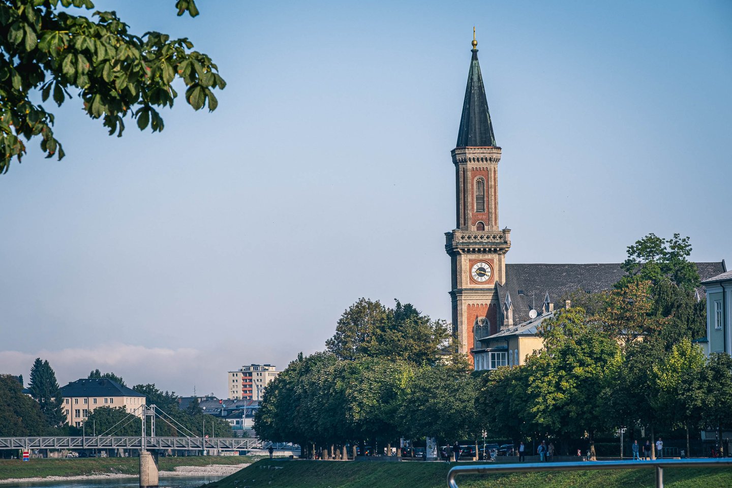 Slender neo-gothic spire of Christuskirche rises above trees along the Salzach river in Salzburg