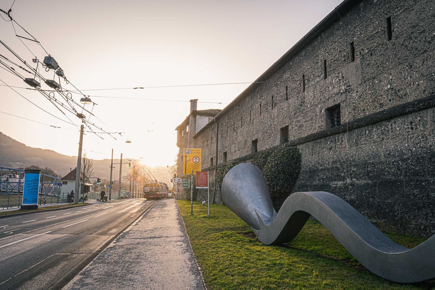 Connection sculpture by Manfred Wakolbinger with wavy metal form beside the old Nonntal bastion wall and tram passing at sunrise in Salzburg