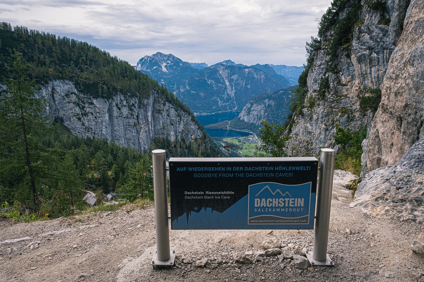 A dark Dachstein Salzkammergut board reading Goodbye from the Dachstein Caves stands on the gravel outside the cave exit, high above the Hallstaetter See in its wooded valley.