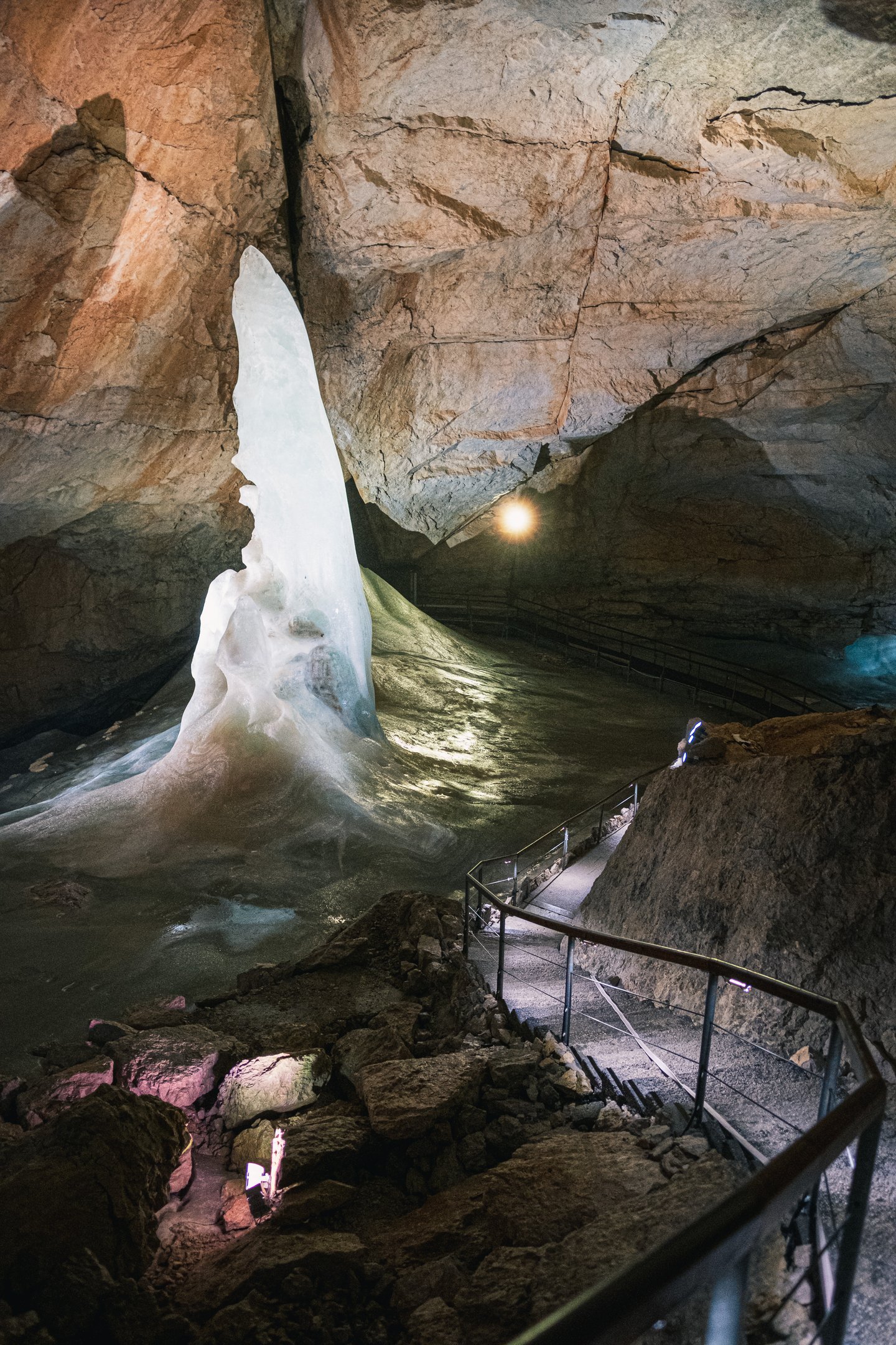 A tall blade of white ice rises from the floor of a warm-lit rock chamber in the Dachstein Giant Ice Cave, seen from the top of the visitor stairs.