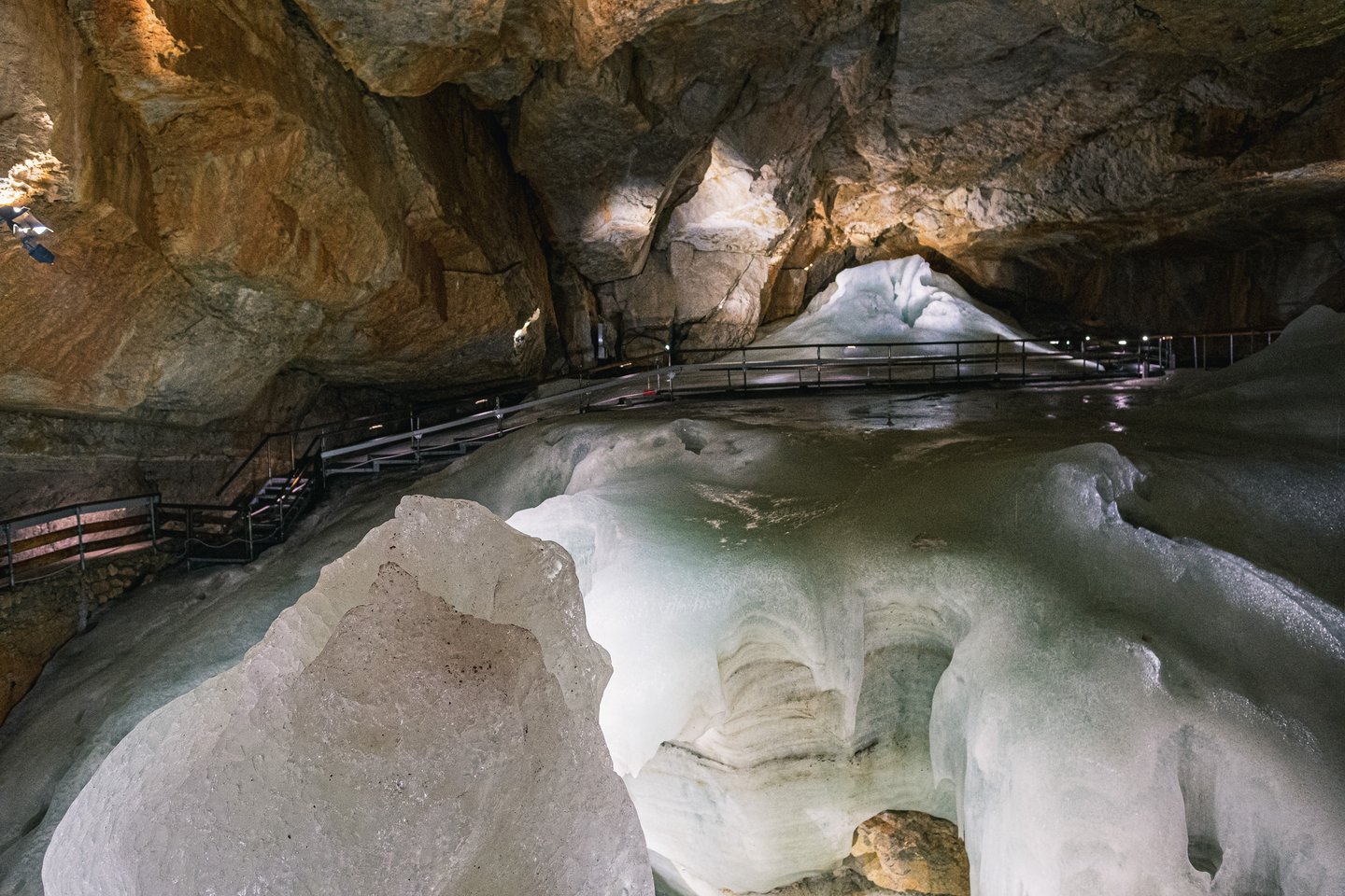 A broad field of layered white ice sweeps across the cave floor, with the metal stairs and railings of the visitor route climbing the rock wall on the left.