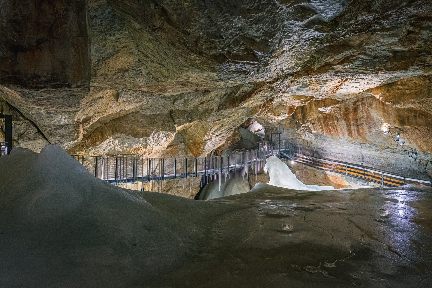 A long steel walkway bridge crosses high above the grey ice floor of a broad chamber in the Dachstein Giant Ice Cave, with icicles hanging from its underside.